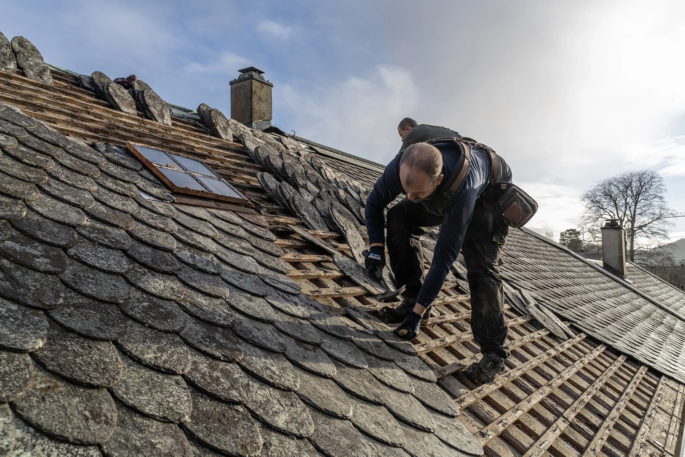 To personer reparerer et tak med gamle takstein, noen av dem er fjernet og andre er fortsatt på plass.