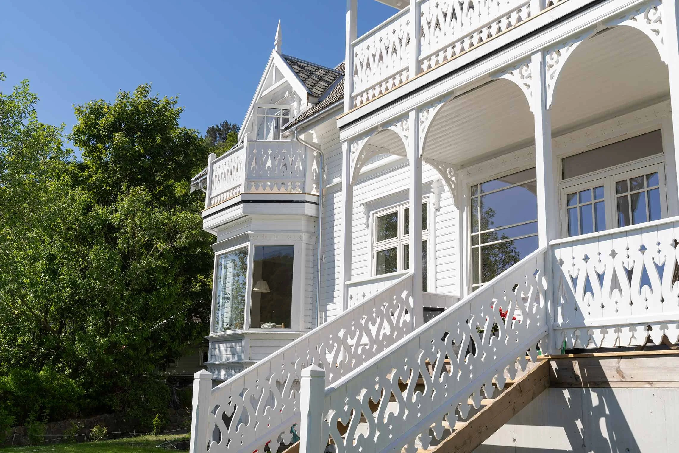 White Victorian-style house with ornate trim and stairs leading up to a porch, surrounded by green trees under a clear blue sky.