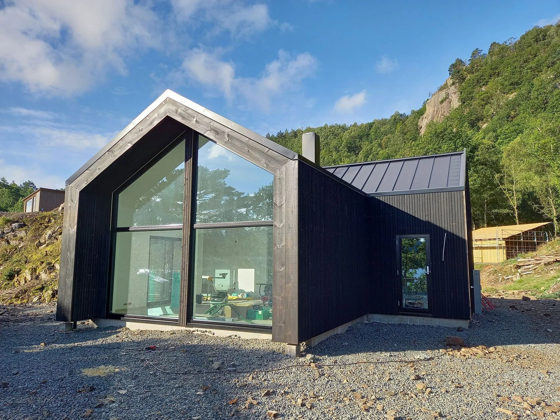 Modern black house with large glass windows and a peaked roof, situated on a gravel lot with a wooded hillside in the background.