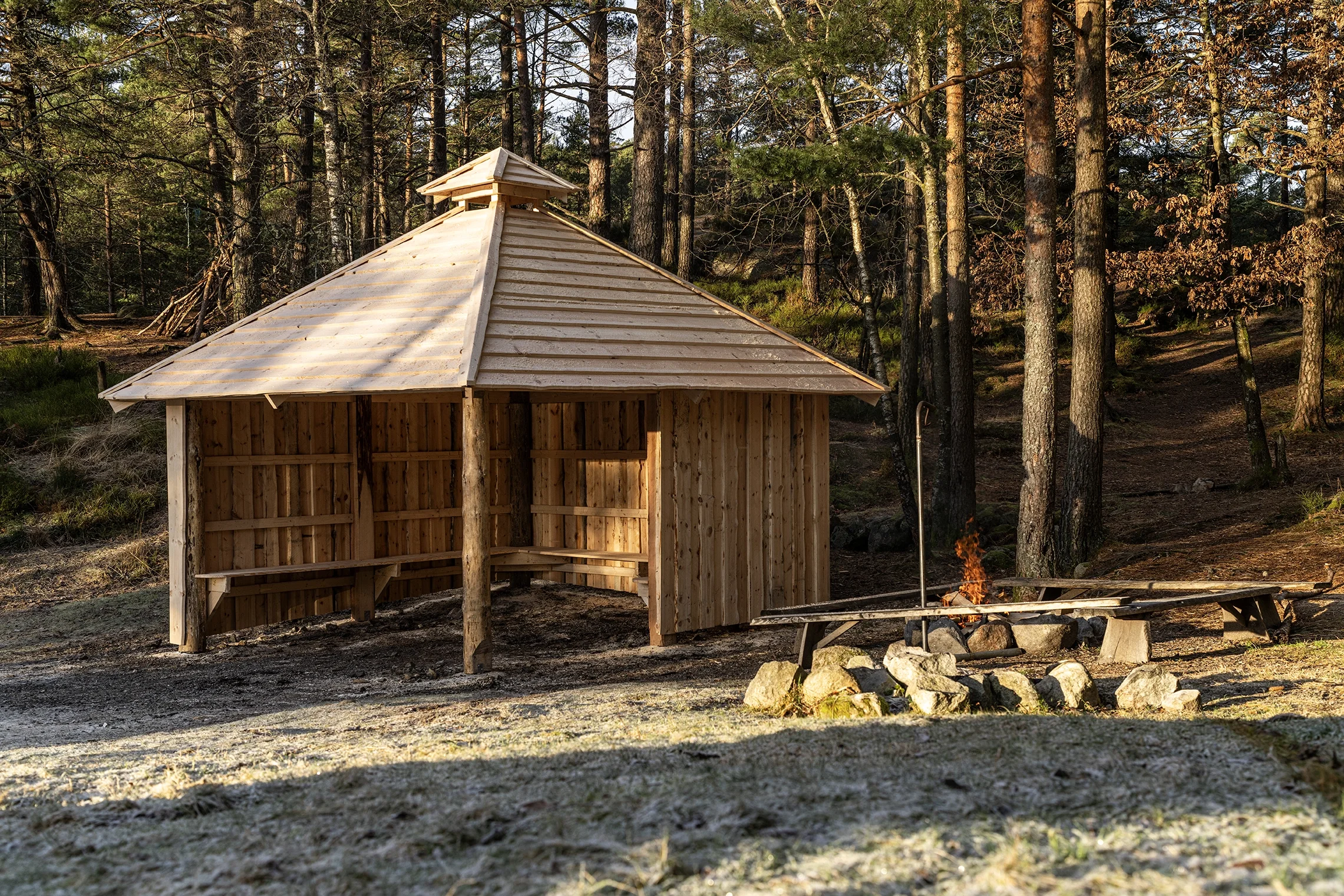 A wooden shelter with a pitched roof in a forested area, with a small fire burning on a makeshift stone and wood fire pit nearby.