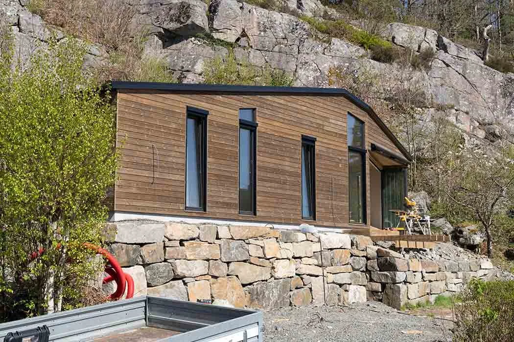 Modern house with wooden siding built on a stone foundation, perched on a hillside, with construction tools on the porch.