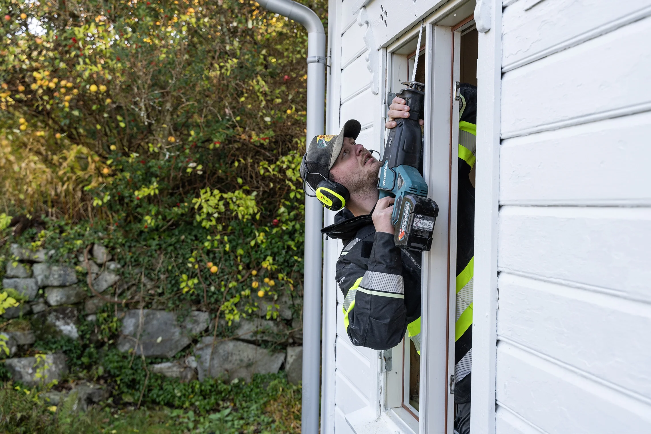 A man wearing a black jacket with reflective stripes and a hat, using a cordless power drill to cut a piece of metal trim on a white house exterior, standing at an open window with a person partially visible behind the wall.