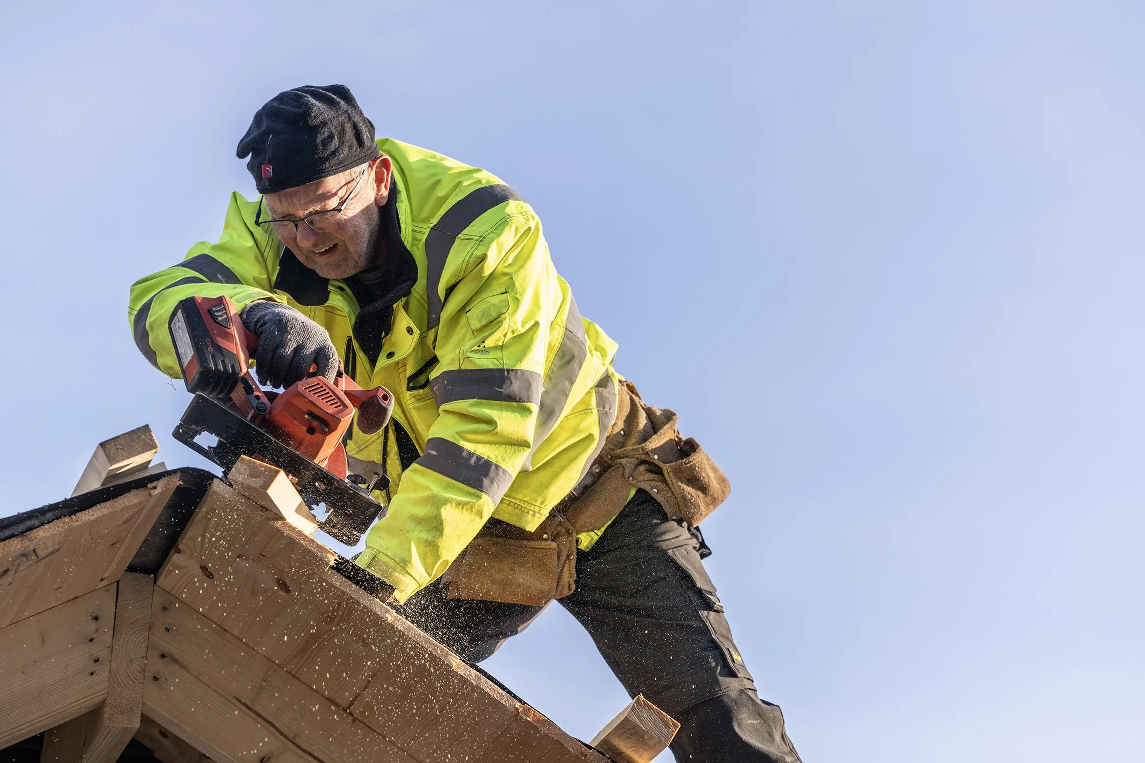 A man in a yellow safety jacket and black beanie using a saw to cut wood on a construction site.