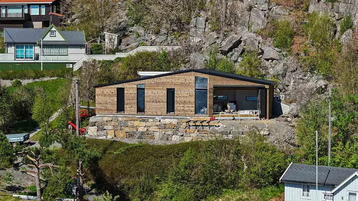 Modern wooden house built into a hillside with large windows, a stone retaining wall, and a forested backdrop.