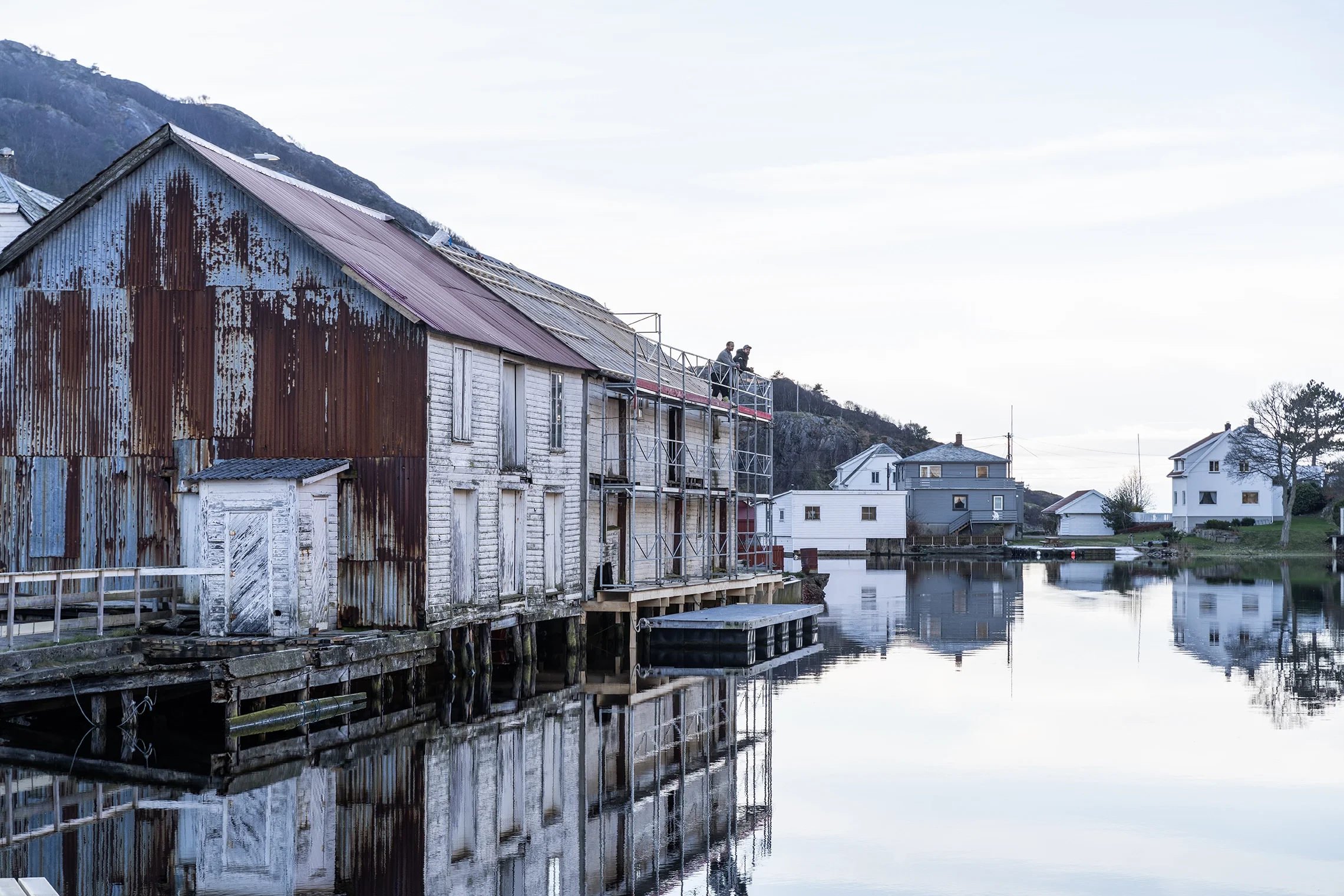Rusty old building beside a calm river with modern houses in the background and a few people on scaffolding at the top of the building.