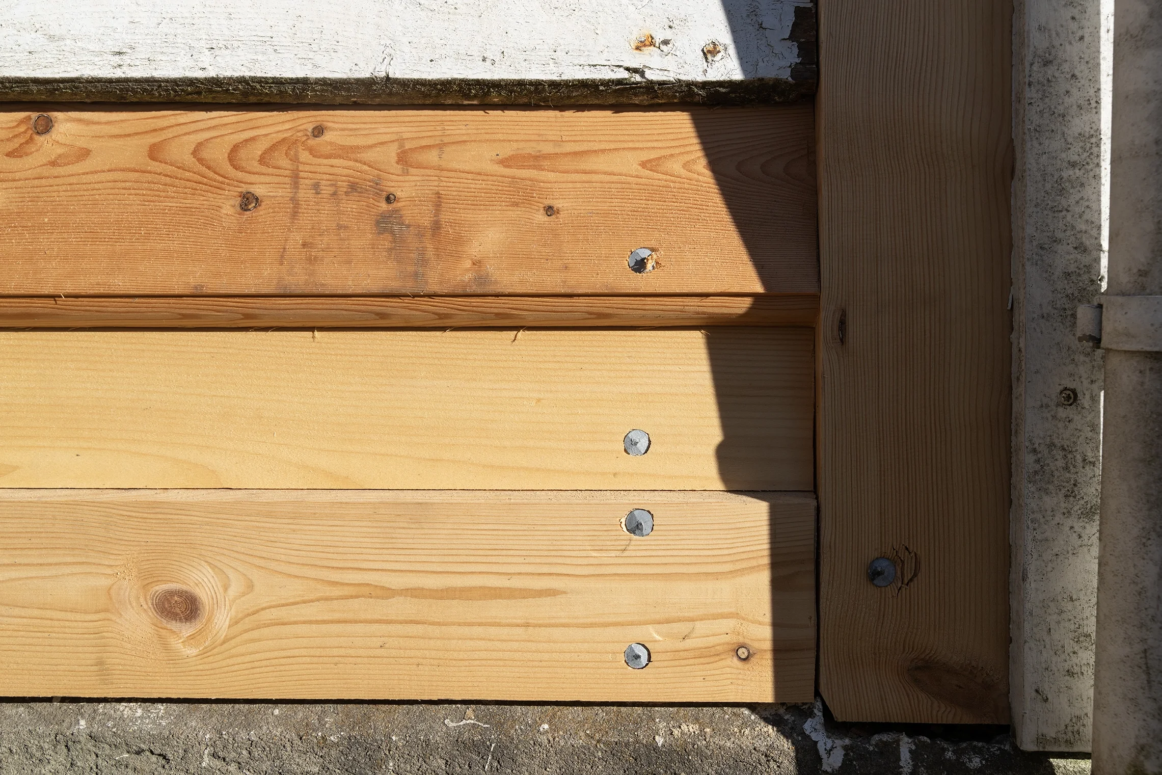 Close-up of wooden boards attached to a concrete surface with screws, with the sunlight casting shadows on the wood.