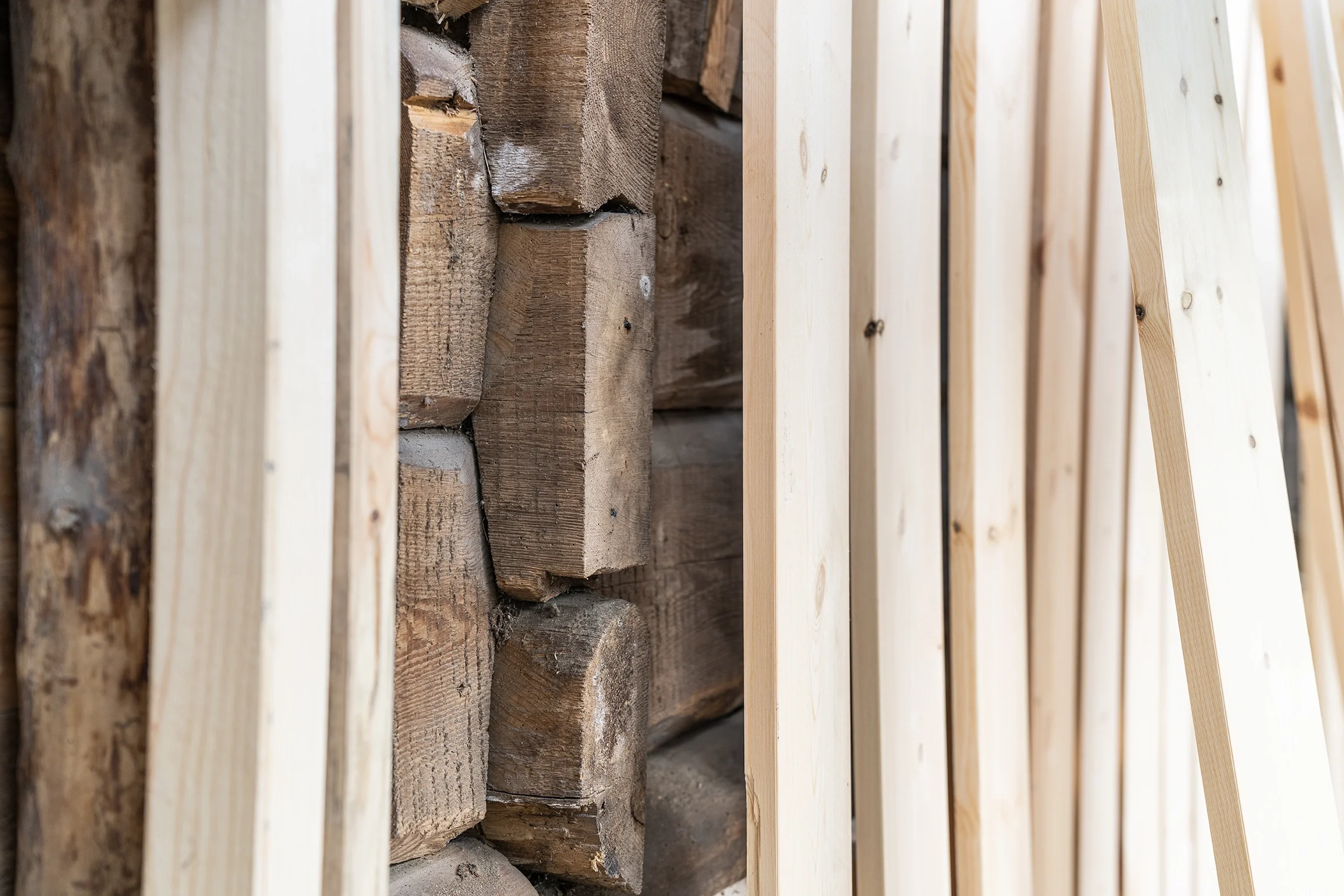 Close-up view of wooden construction materials, showing stacks of rough-cut logs on the left and light-colored finished wood planks on the right.