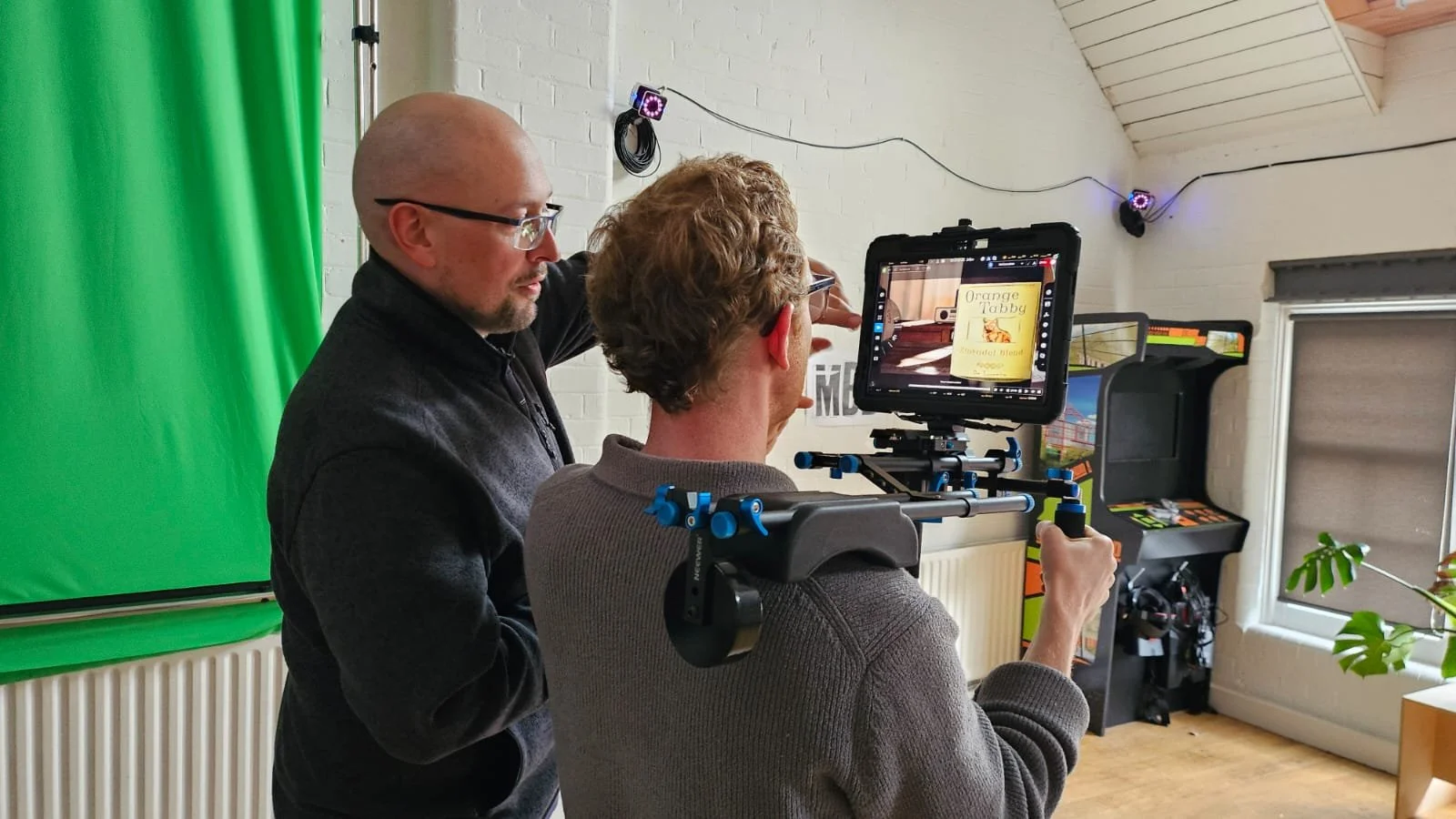 Two men filming indoors, one with a camera rig and an external monitor, with a green screen on the left and arcade games on the right.