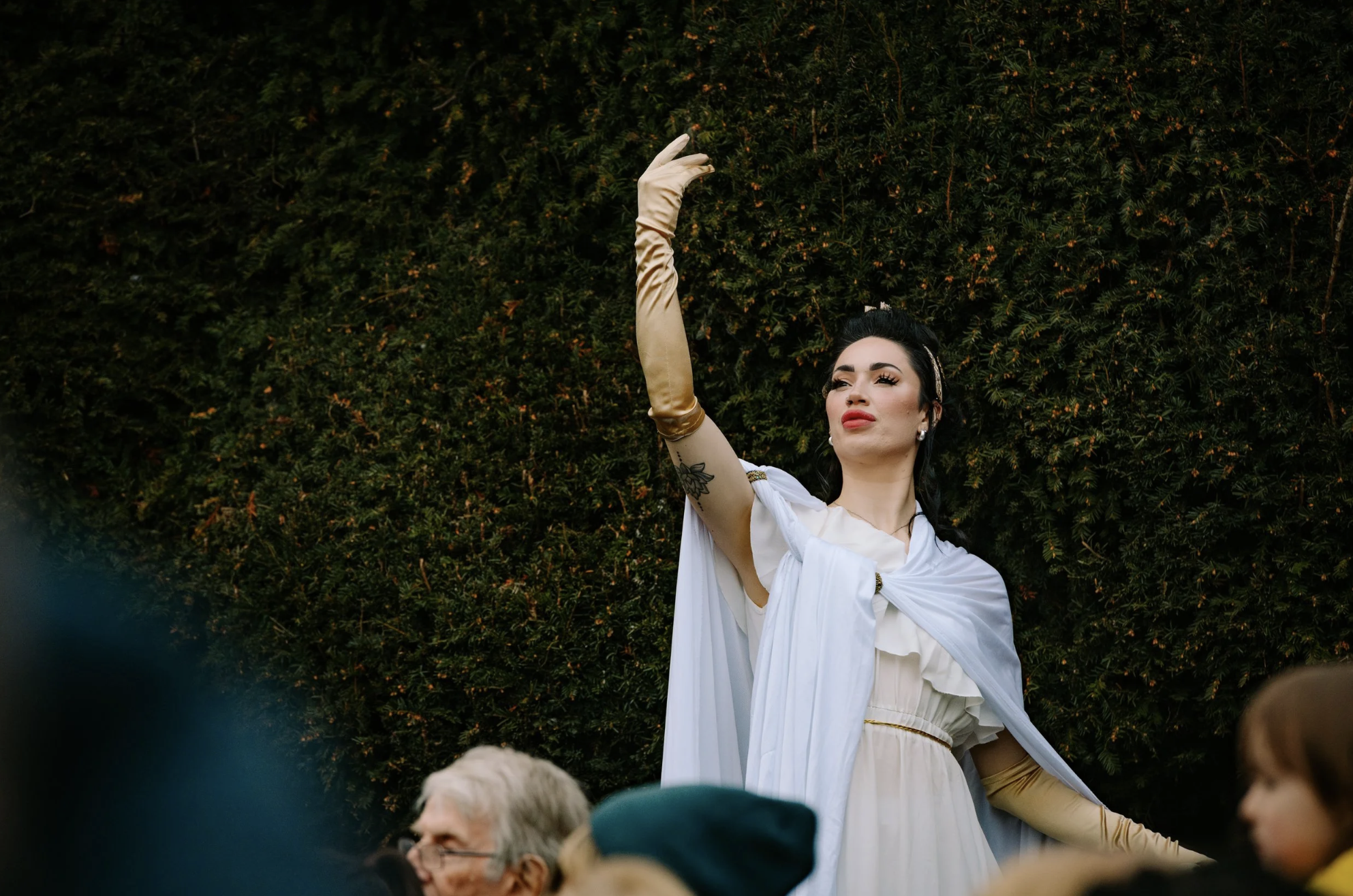 Female actress stood in front of hedge with her arm raised to the sky