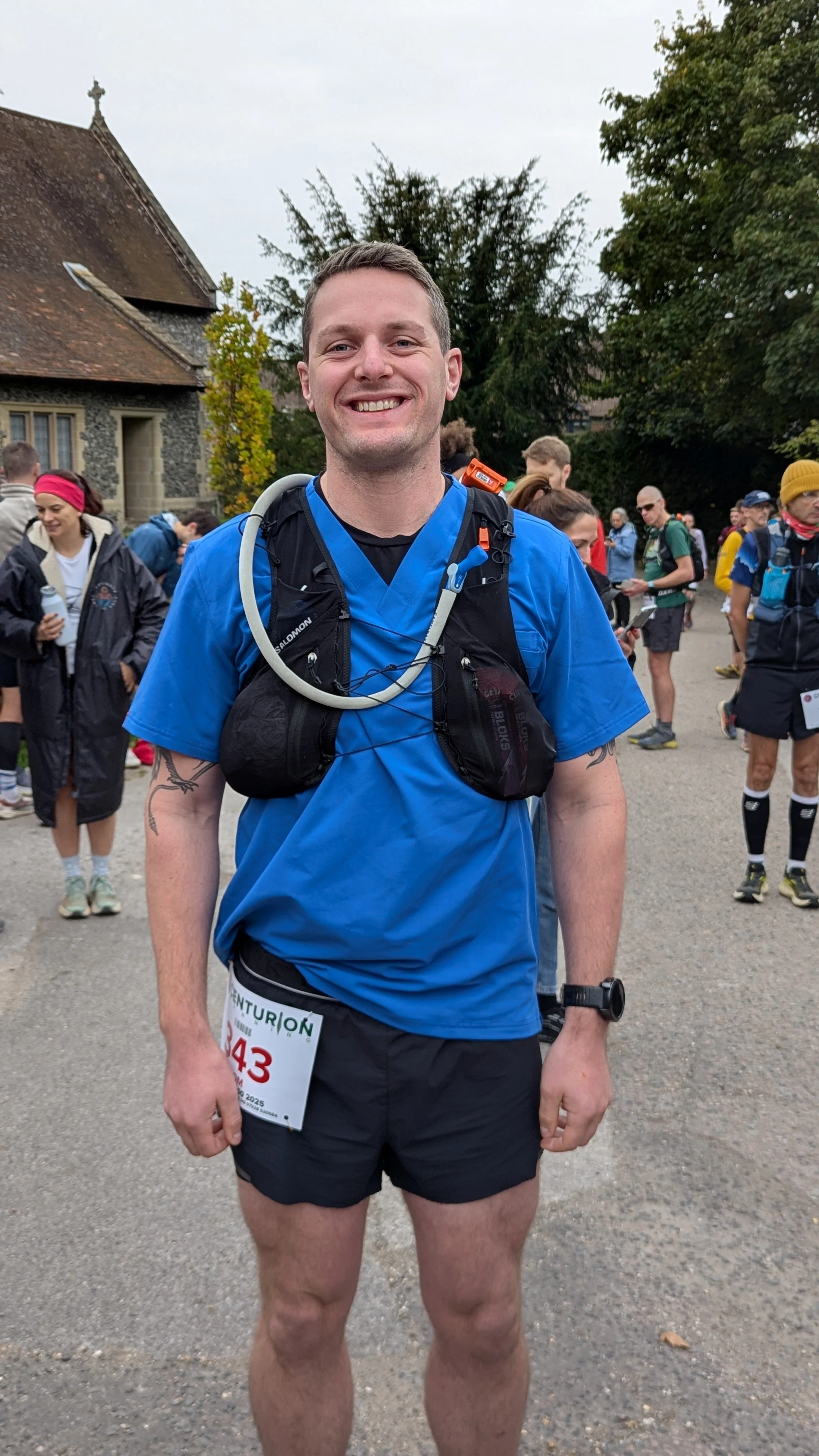 A smiling man wearing a blue running shirt, black shorts, and a race bib, standing among a group of runners outdoors with trees and old buildings in the background.