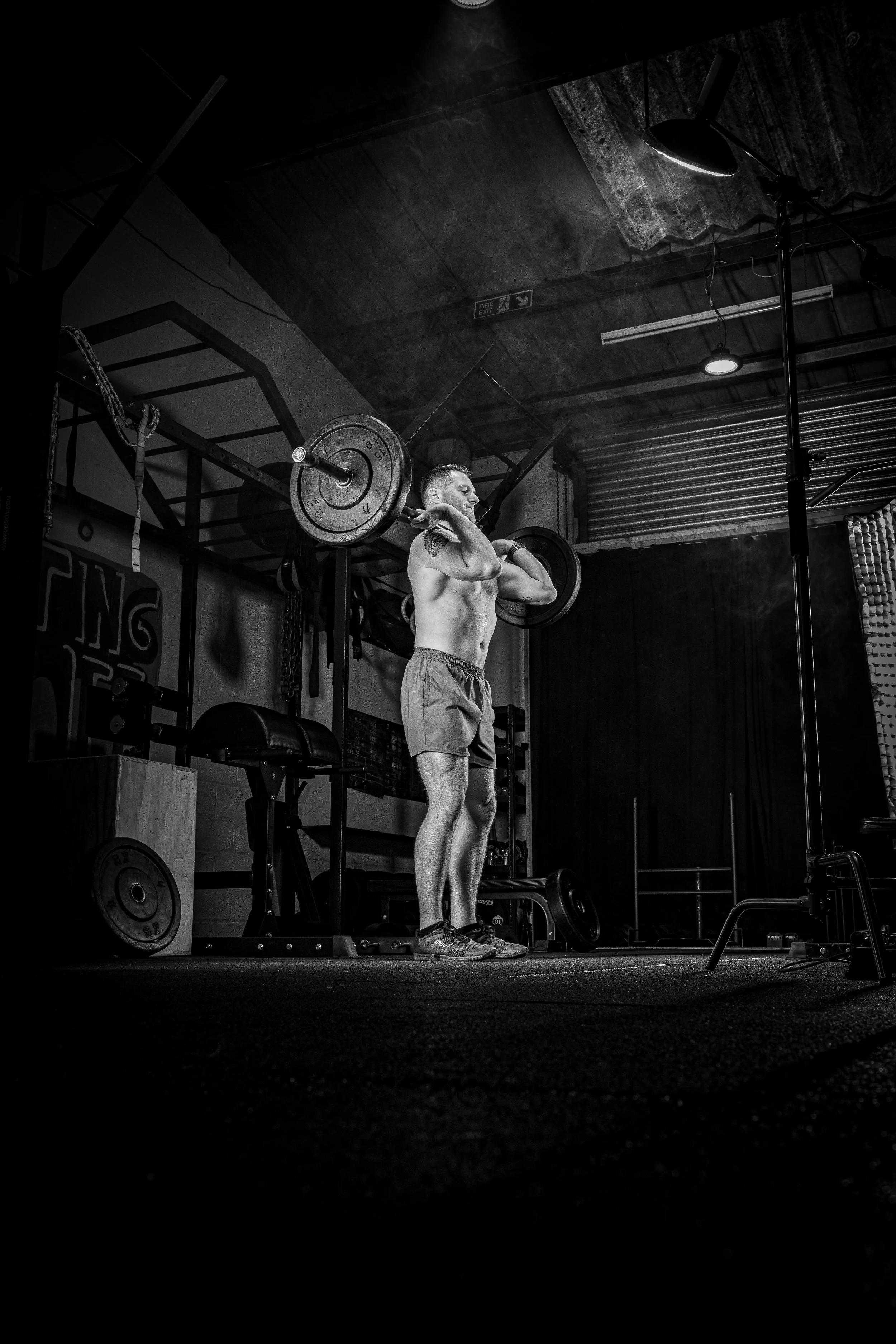 A shirtless man lifting a barbell with weights in a gym, standing on a rubber mat in a black-and-white photo.