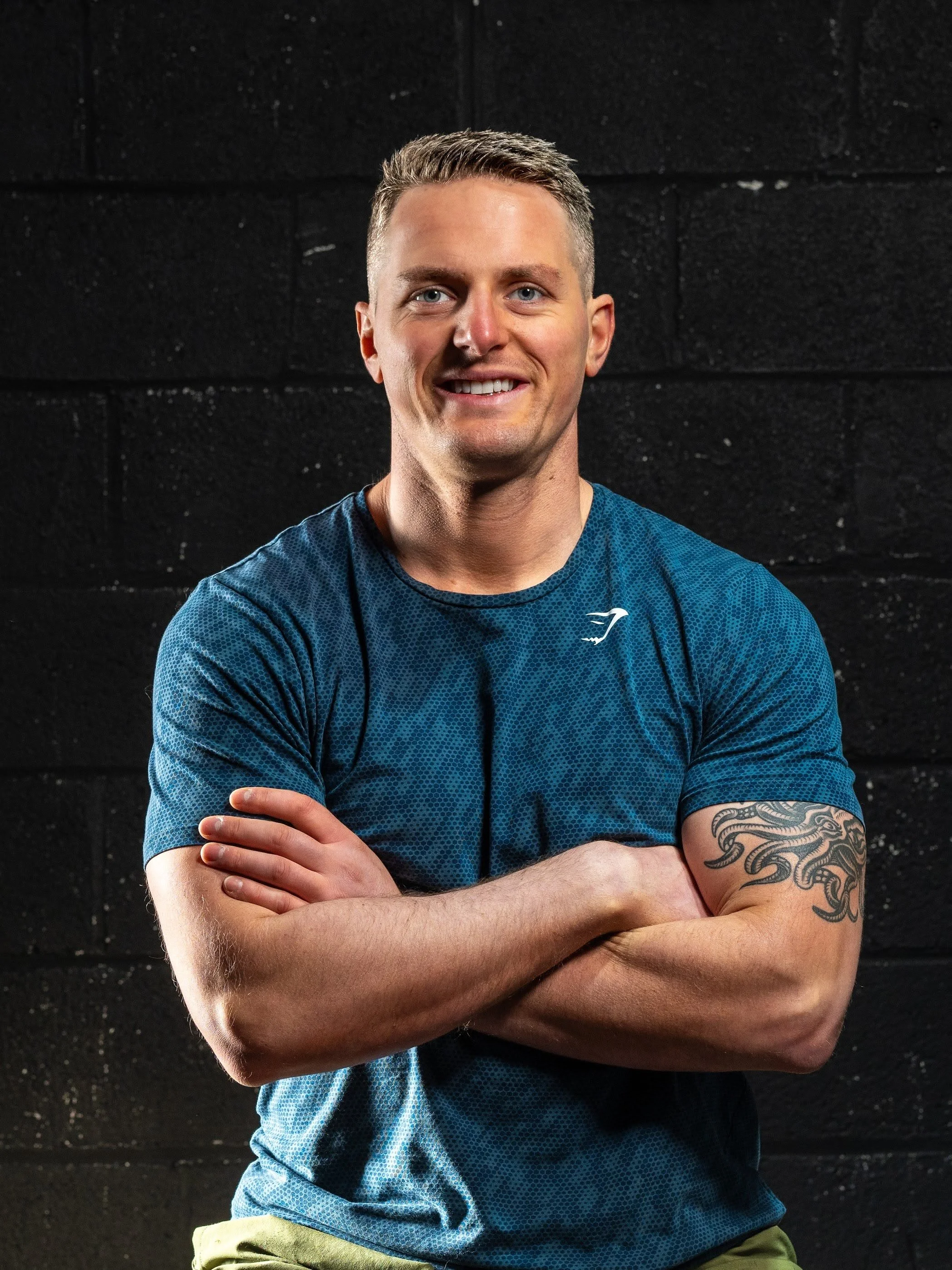 A man with short hair and a tattoo on his right arm, wearing a blue athletic shirt, smiling with arms crossed, standing in front of a black wall.
