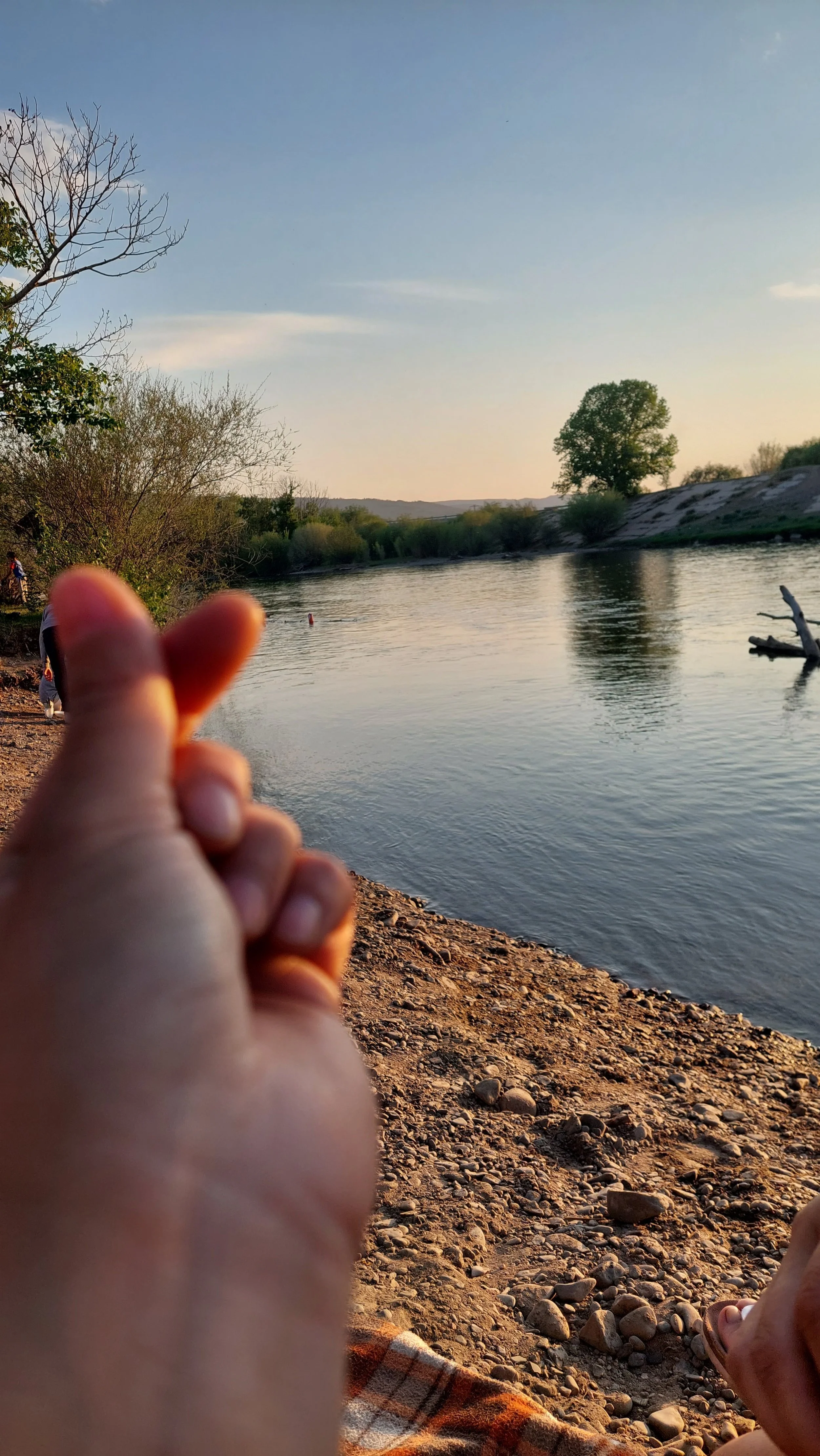 Person near a river with a hand making a finger heart gesture in the foreground, trees lining the riverbank, calm water, and a clear sky at sunset.