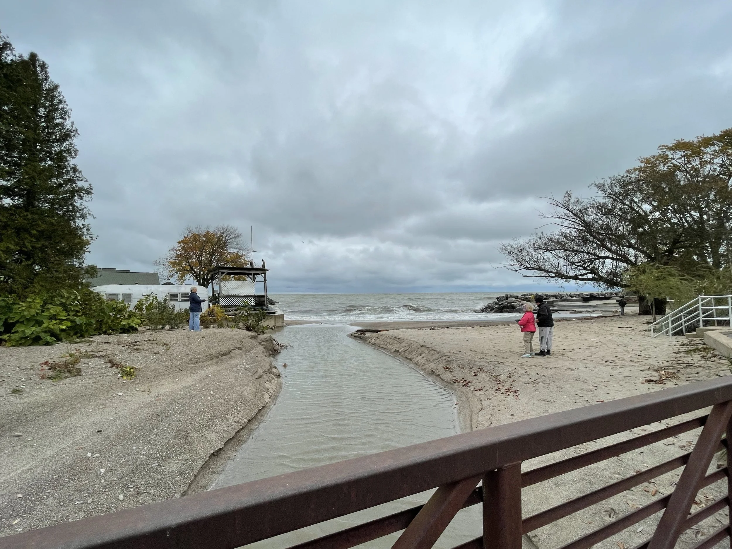A cloudy day at a beach with a small stream flowing into the ocean. There are three people: one near the stream, and two walking on the sand. Trees and a rock jetty are visible in the background.
