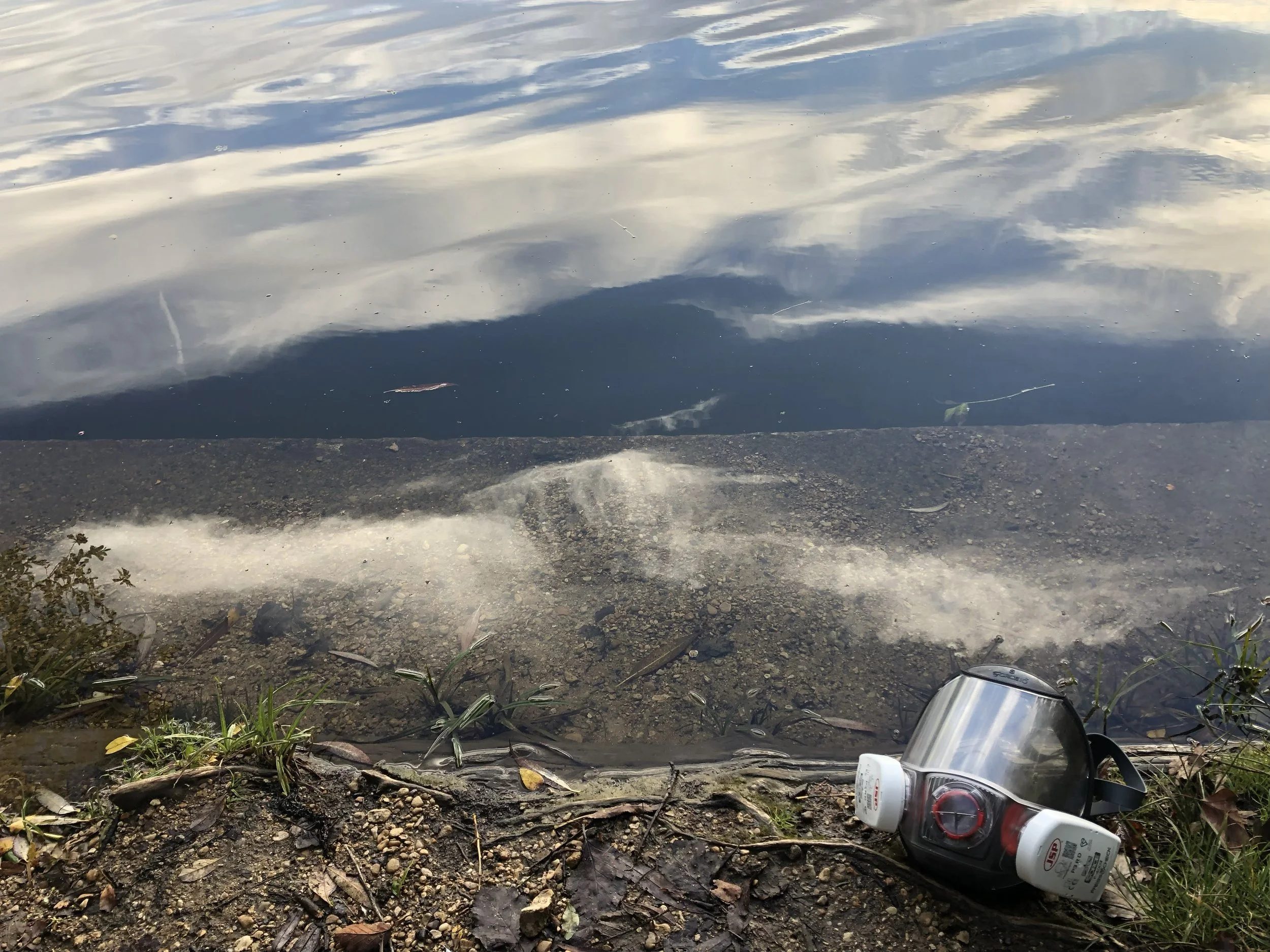 A face shield, medical mask, and a bottle of hand sanitizer on the ground near a riverbank, with the river water reflecting the sky and clouds.