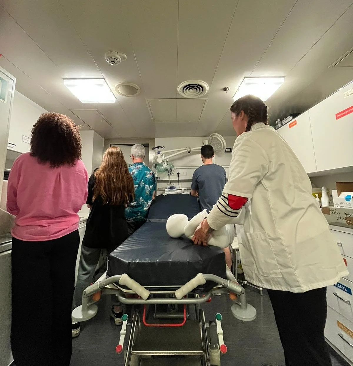 Medical training or demonstration with a mannequin in a clinical room, five clinical staff and a doctor or instructor.