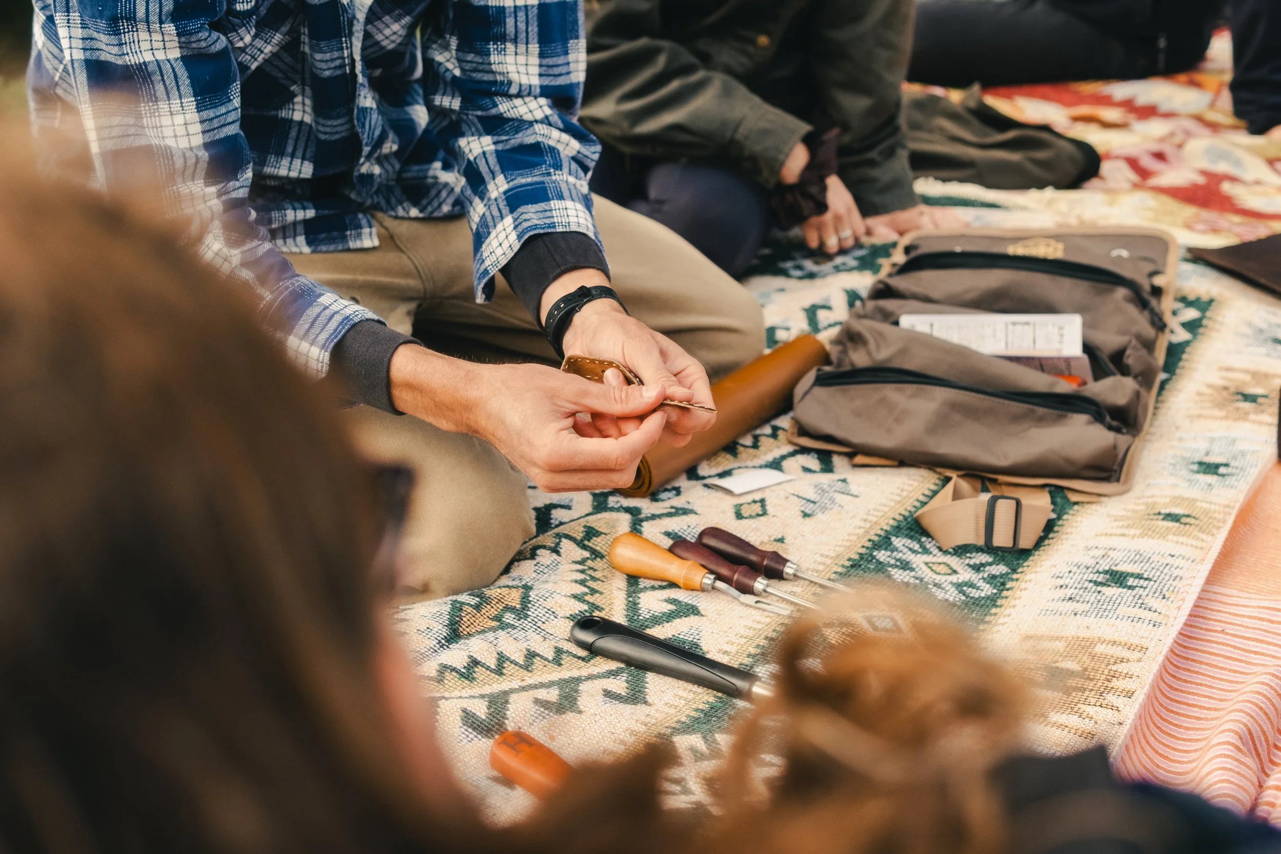 Person in a blue plaid shirt holding a coin, kneeling on a patterned rug with tools and a backpack nearby, with other people sitting on the rug.