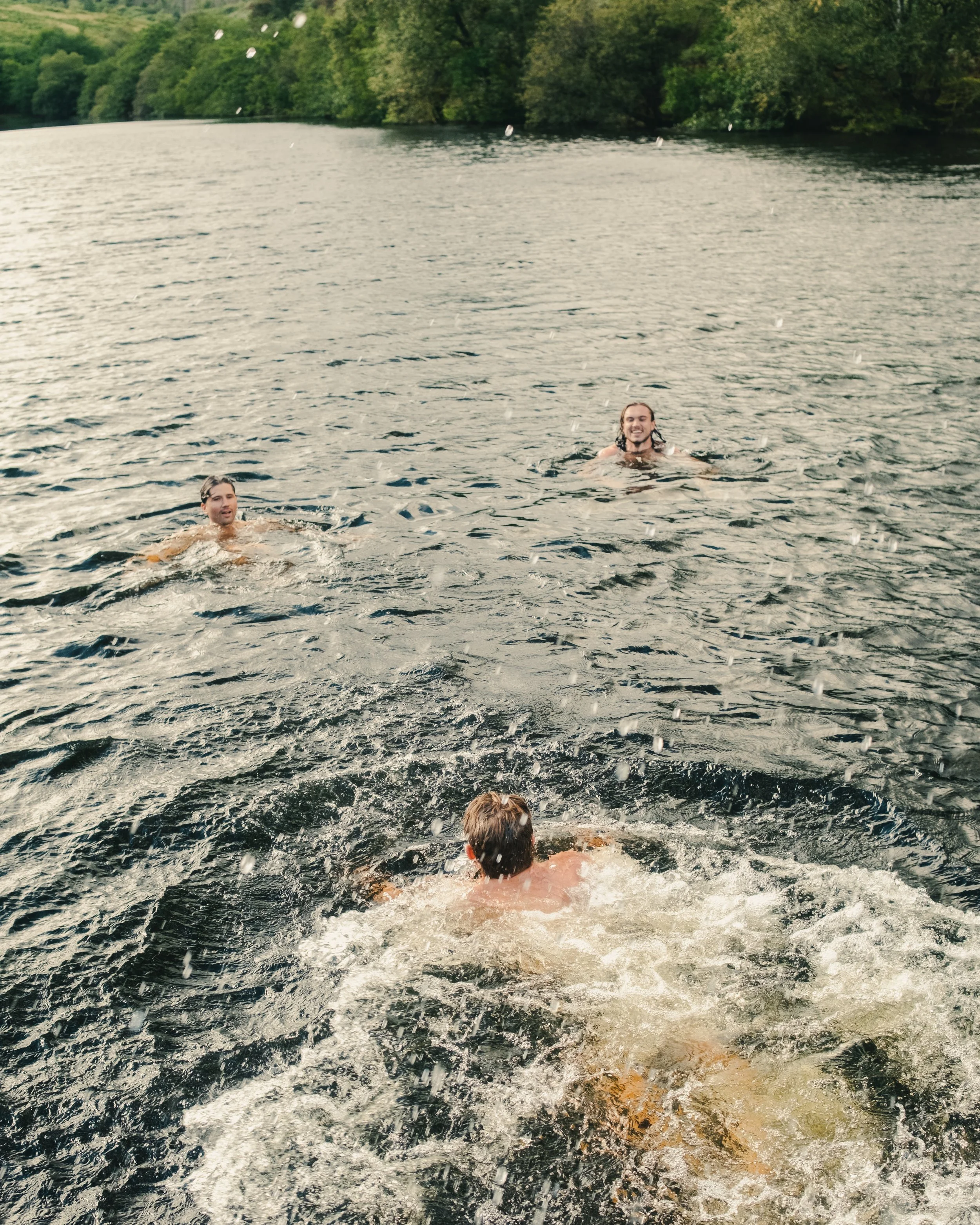 Three young men swimming and floating in a river with trees along the riverbank in the background.