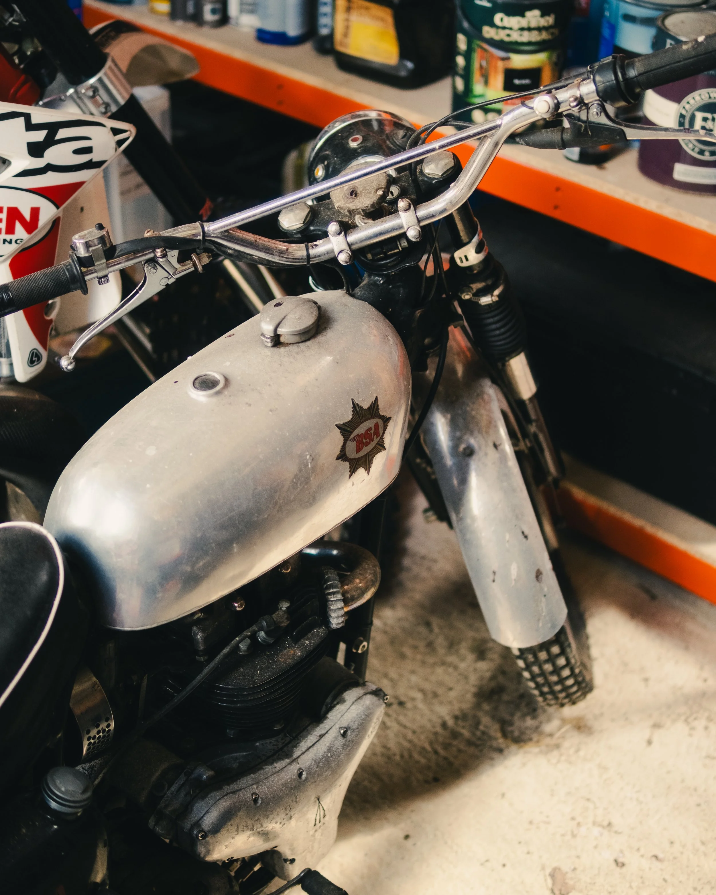 Close-up of a vintage motorcycle with a silver fuel tank and a black and white 'BSA' badge, parked in a garage with tools and shelves in the background.