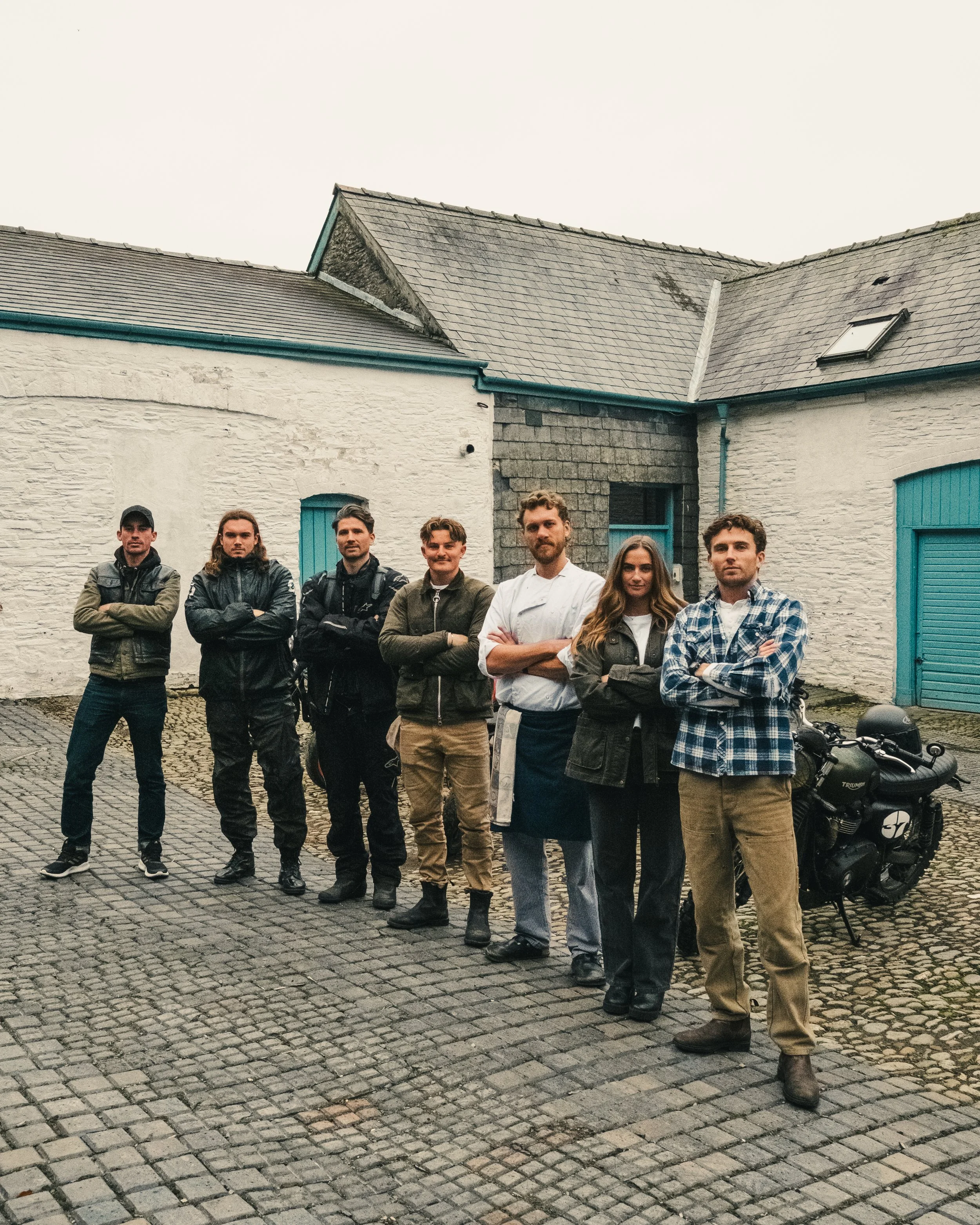 Group of seven people standing outdoors on cobblestone pavement in front of white and gray buildings, some with blue shutters, with a motorcycle parked on the right.