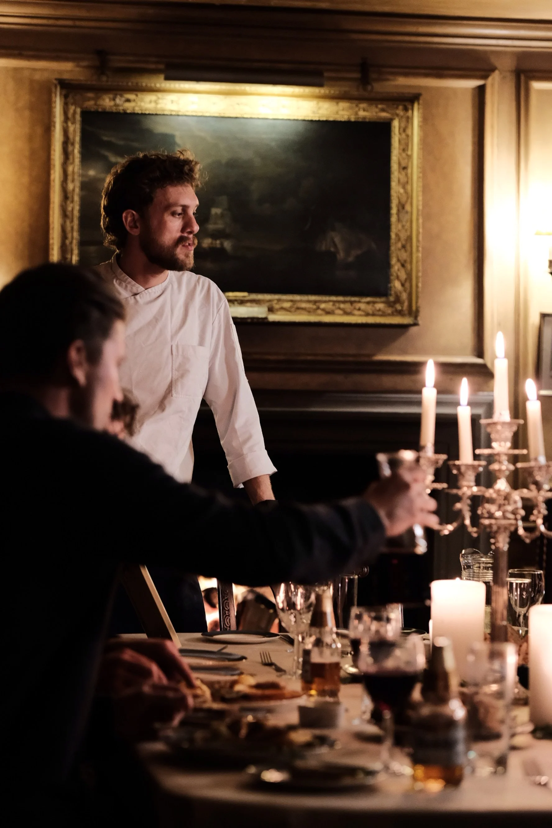 A man in a white shirt standing at a dining table with other seated people, surrounded by lit candles and glasses, in a dimly lit room with a painting on the wall.