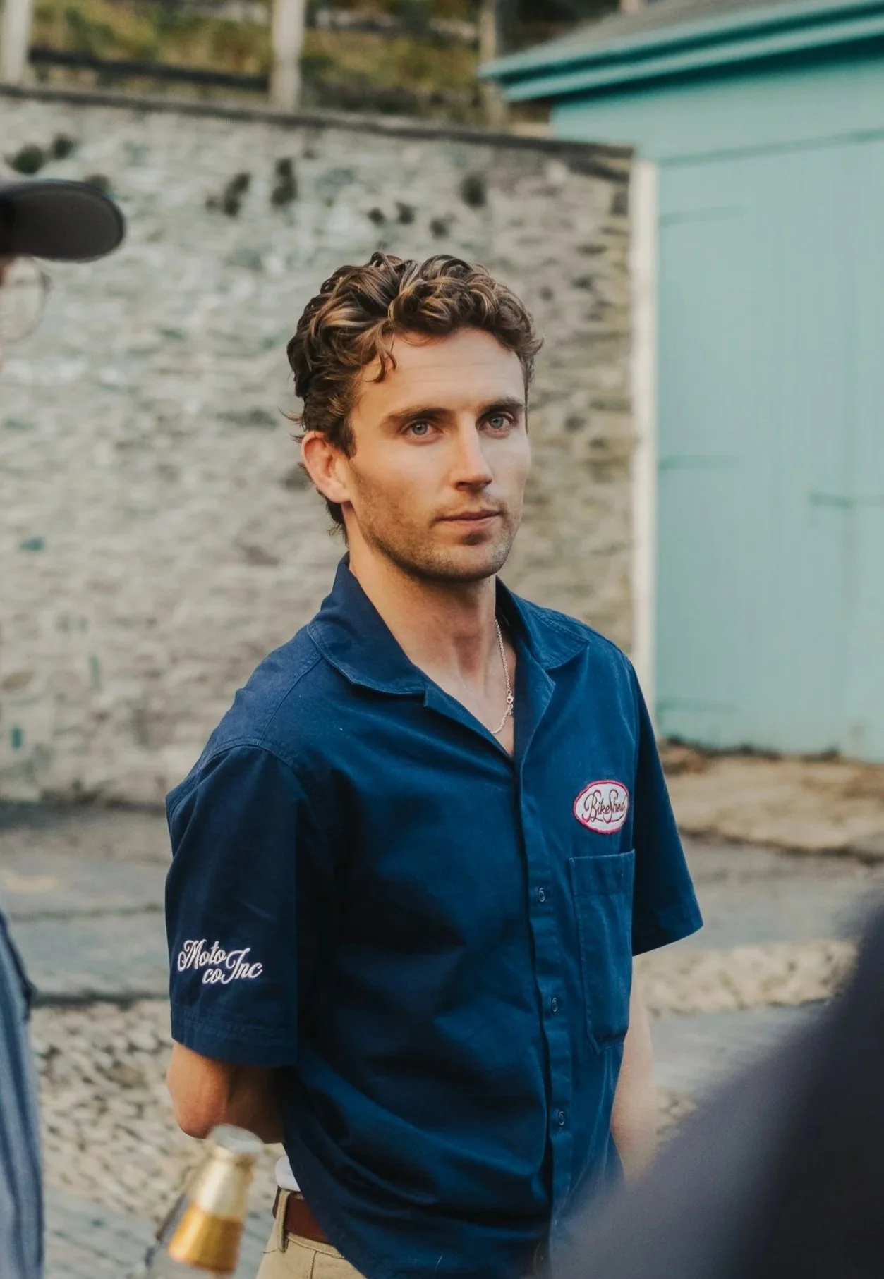 A young man with wavy brown hair and light skin, wearing a dark blue short-sleeved shirt with embroidered patches, standing outdoors with a brick wall and a light blue building in the background.
