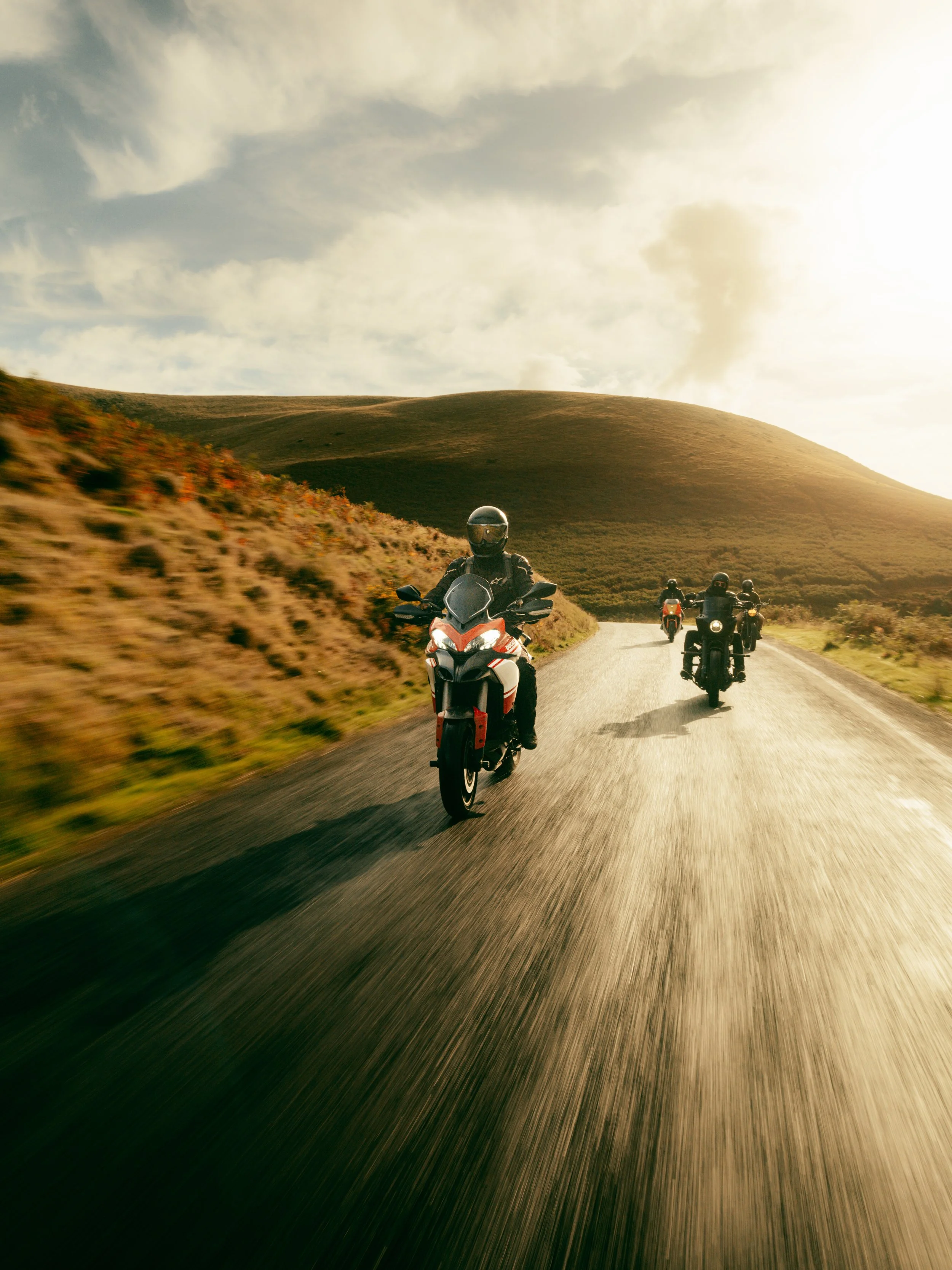 Four motorcyclists riding on a scenic road through a hilly landscape during sunset.