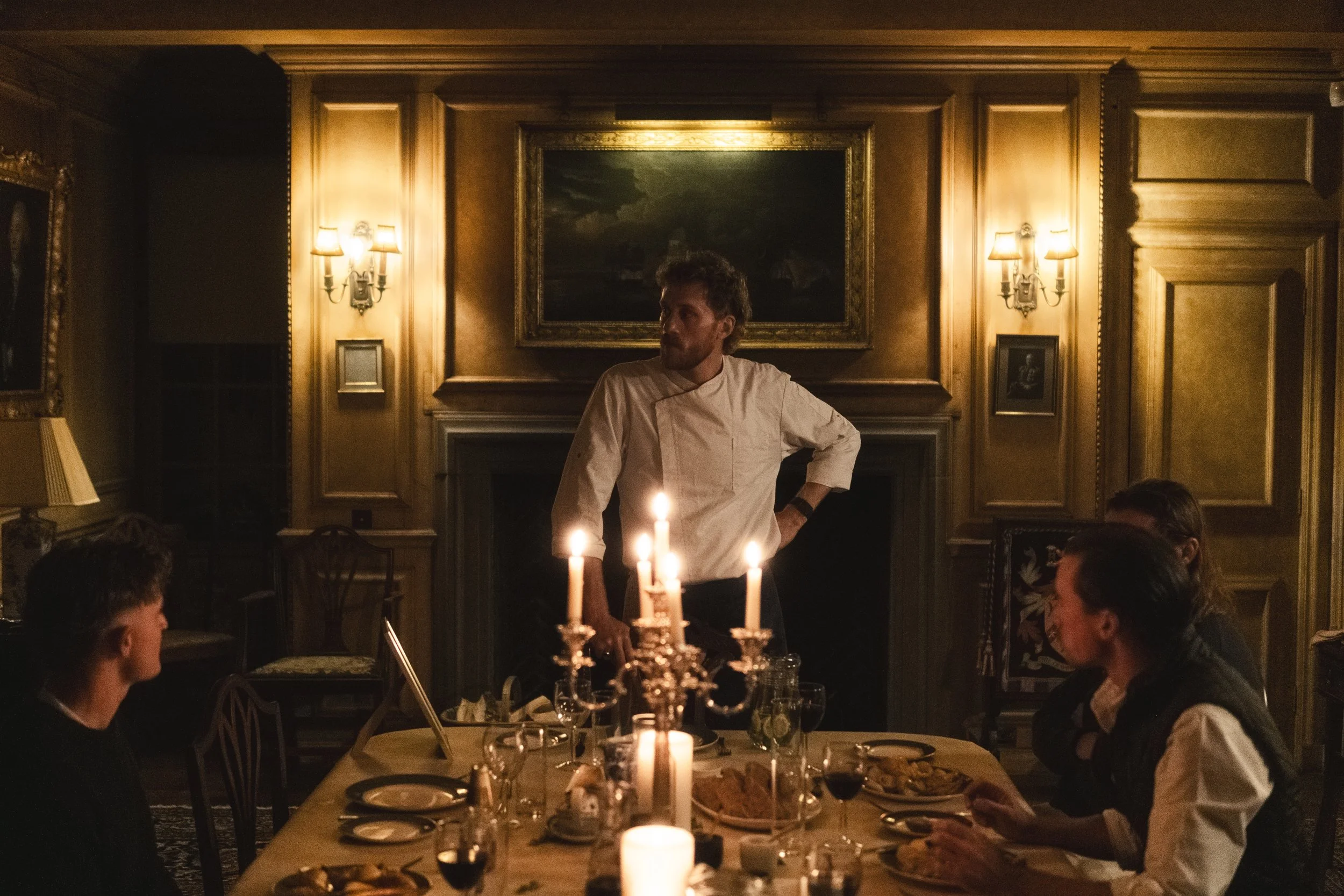 A chef standing at a dinner table in a dimly lit, elegant room with wood-paneled walls, surrounded by four seated guests, with candles on the table.