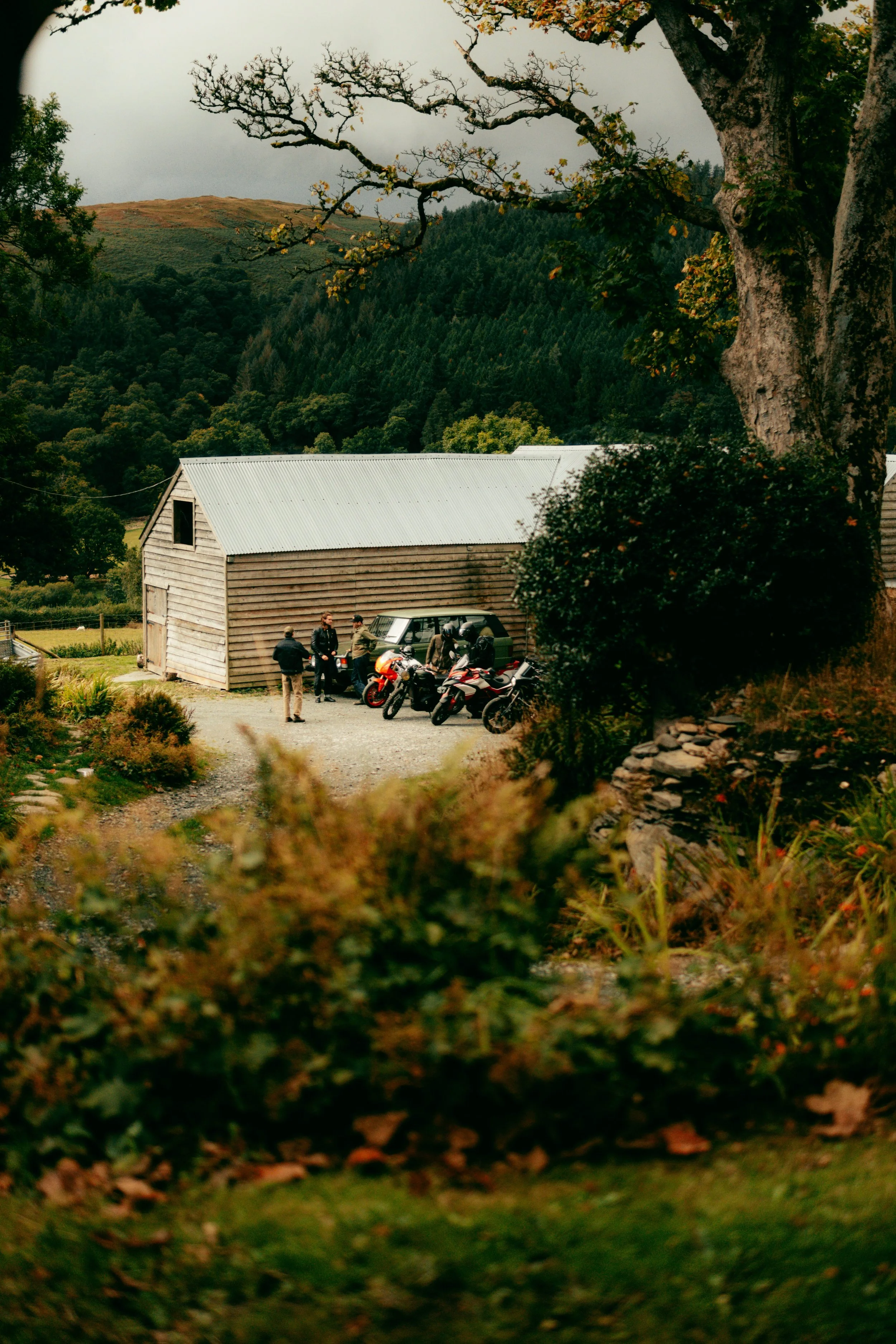 A group of people gathered outside a wooden barn with motorcycles and a van, surrounded by trees and hilly landscape.