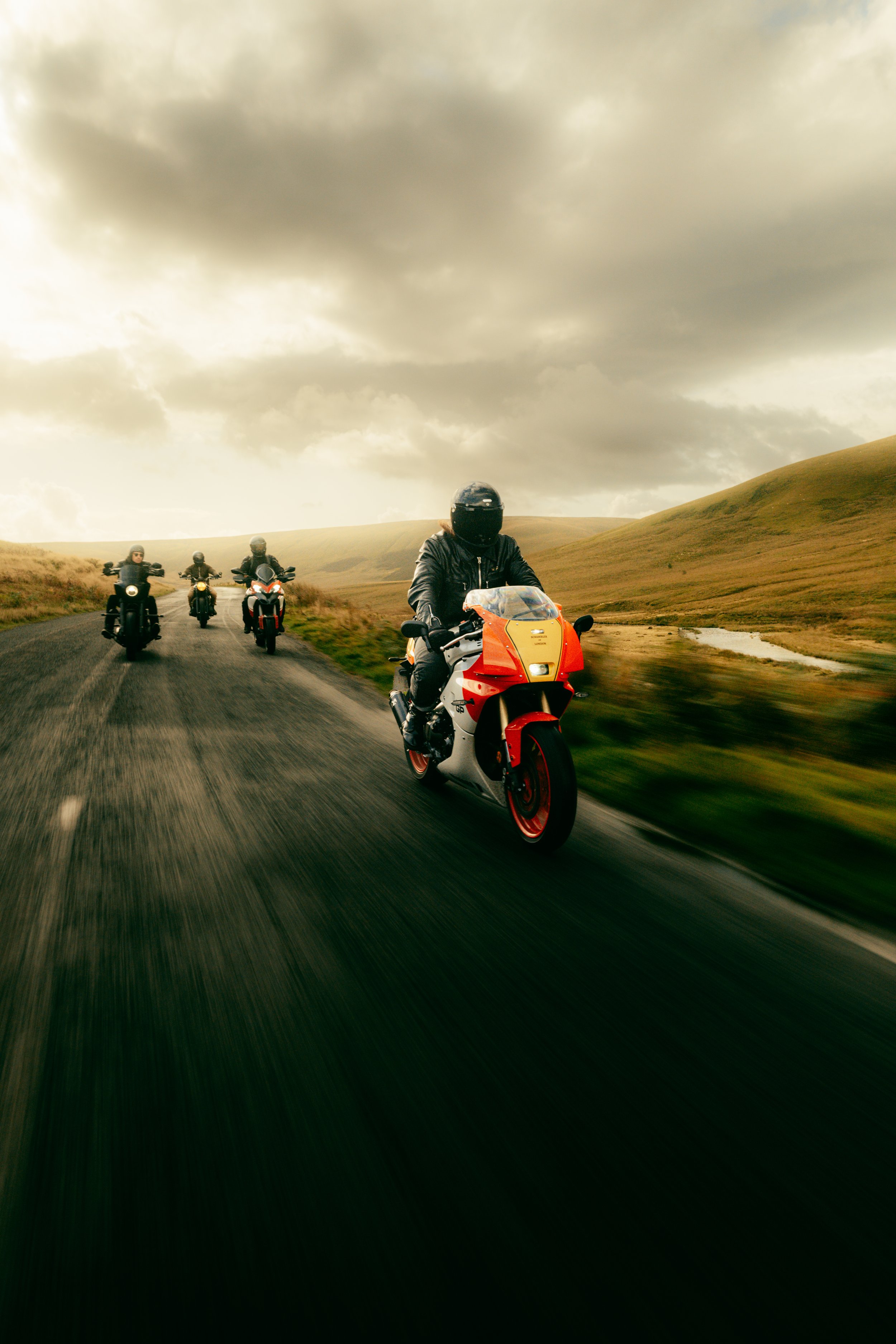 Four motorcyclists riding on a winding rural road through grassy hills under cloudy skies.