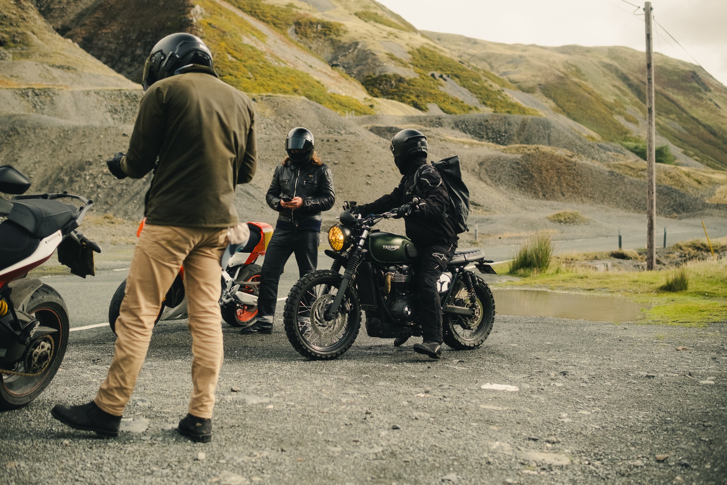 Three motorcyclists with helmets and riding gear standing beside and on their bikes on a mountain roadside.