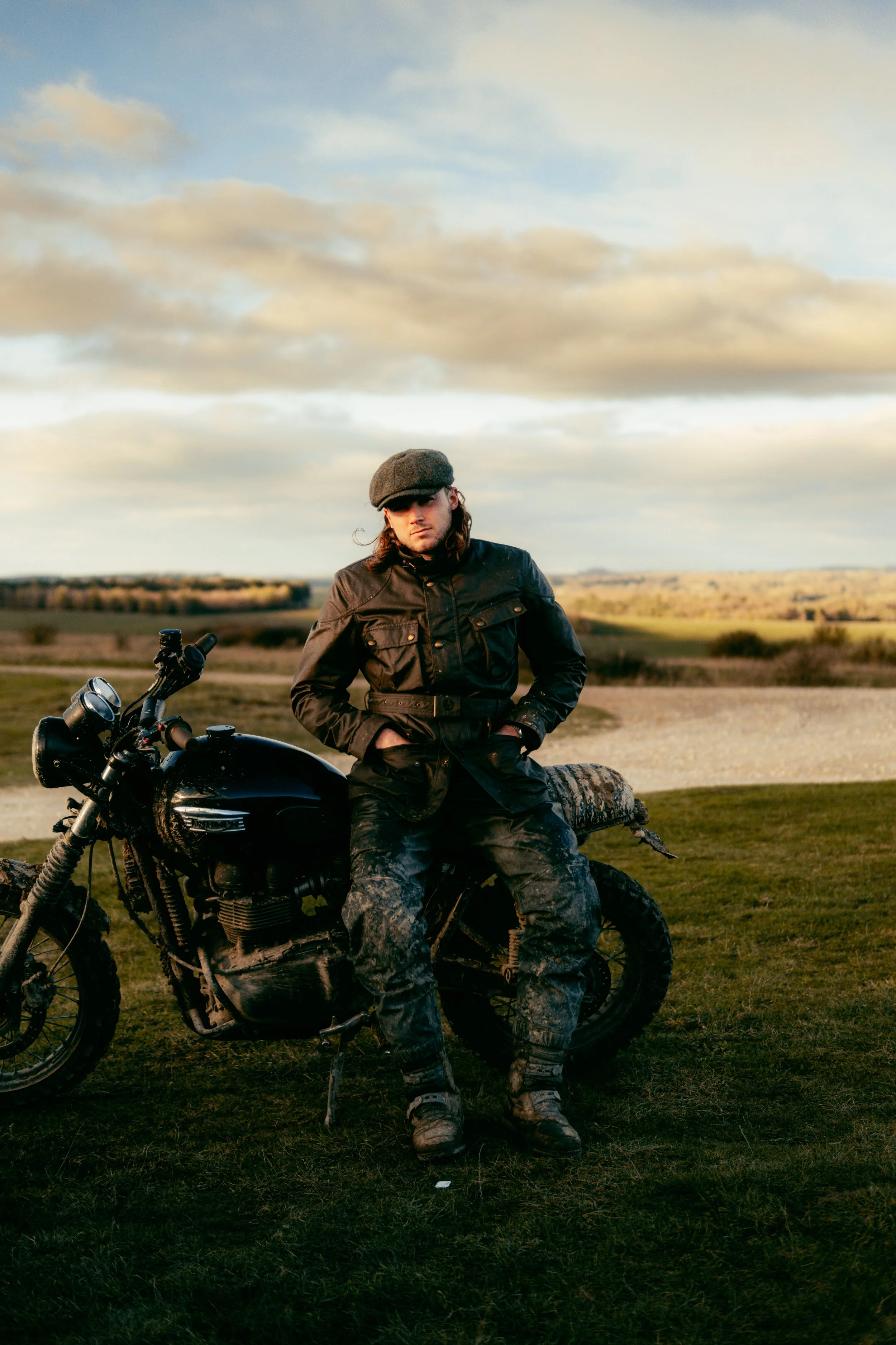 A person sitting on a black motorcycle outdoors in a grassy field during sunset, wearing a black jacket, camouflage pants, boots, and a flat cap.