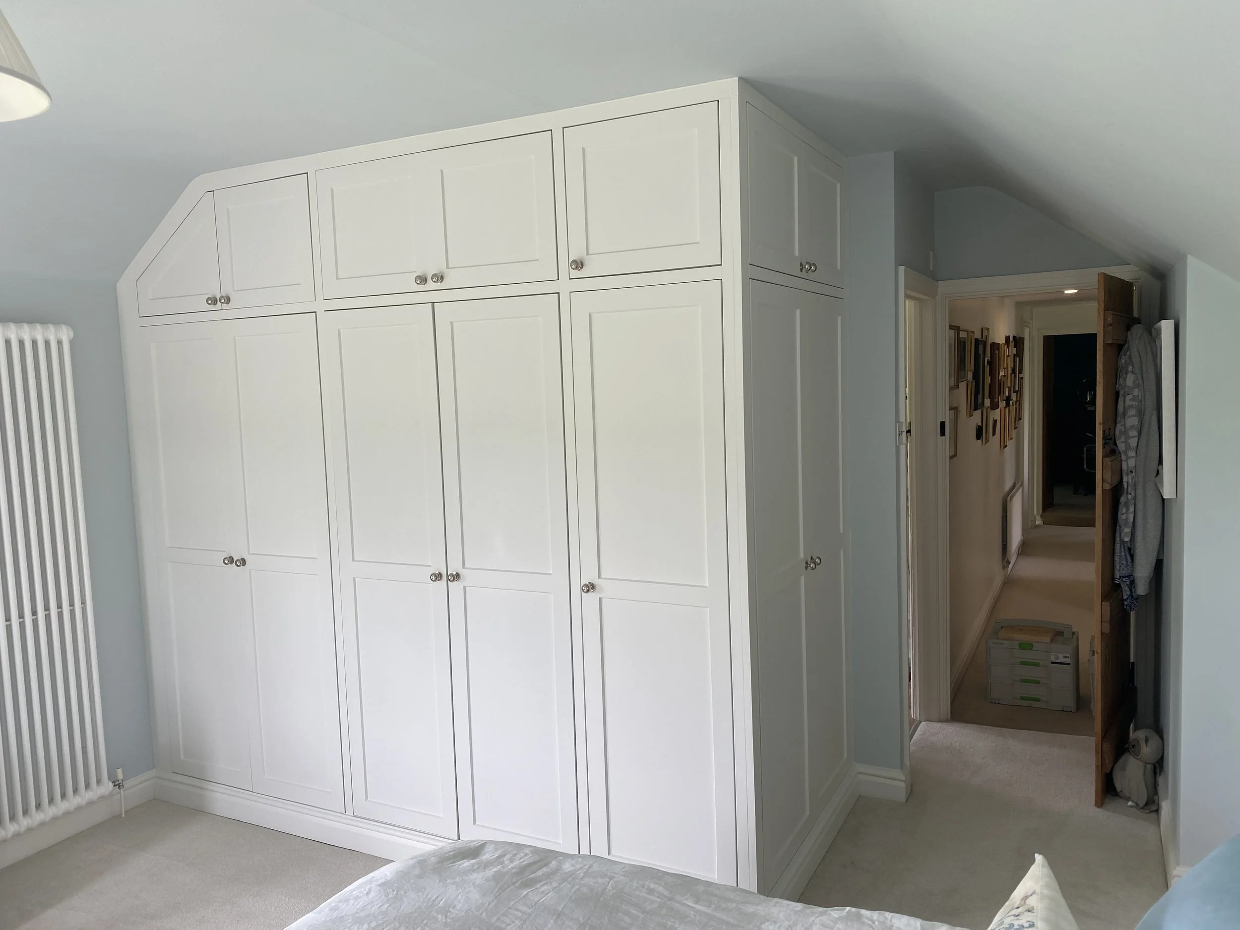 Bright bedroom with white built-in wardrobes, a white radiator, and an open wooden door leading to a hallway decorated with framed artwork.