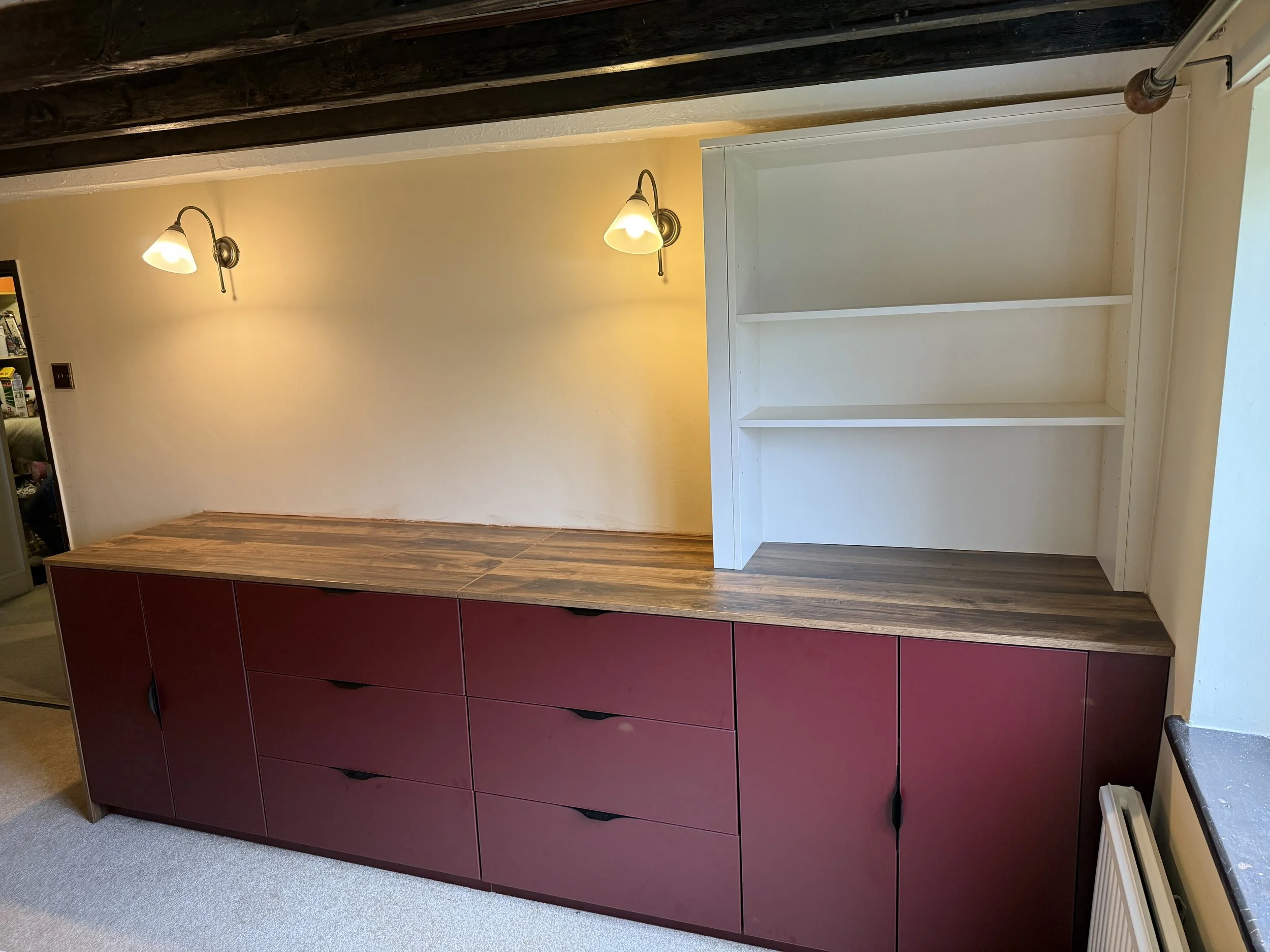 A wooden sideboard with closed cabinet doors and drawers beneath a white empty bookshelf. Two wall-mounted lamps above illuminate the area, with a sloped ceiling and dark exposed beams overhead.