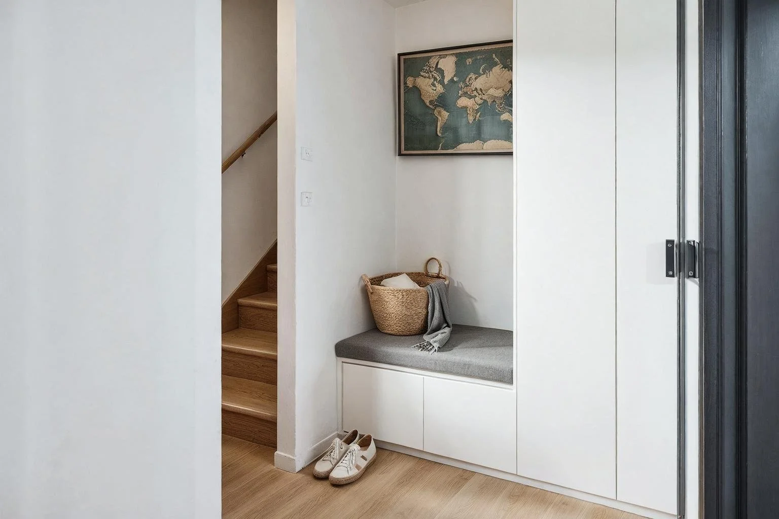 Interior of a modern home entryway with a wooden staircase, framed world map on the wall, a built-in bench with a gray cushion, a wicker basket with a blanket and pillow, and a pair of white sneakers on the light wood floor.
