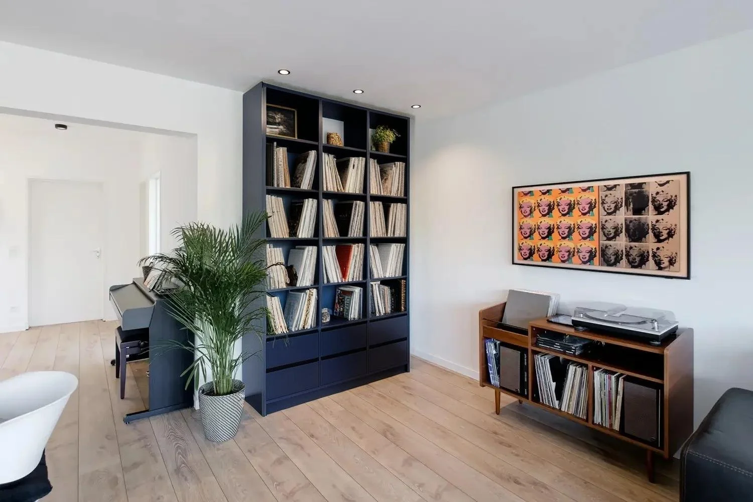 Living room with a tall black bookshelf filled with vinyl records, a potted plant, a wooden sideboard with a record player and vinyl collection, modern artwork on the wall, and hardwood floors.