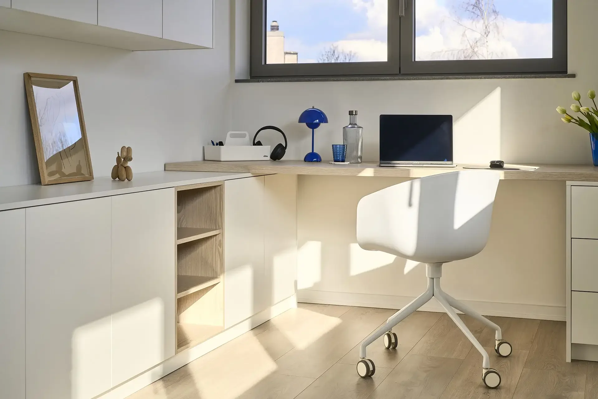 Bright minimalist home office with white desk, rolling white chair, and large window showing a blue sky and trees.