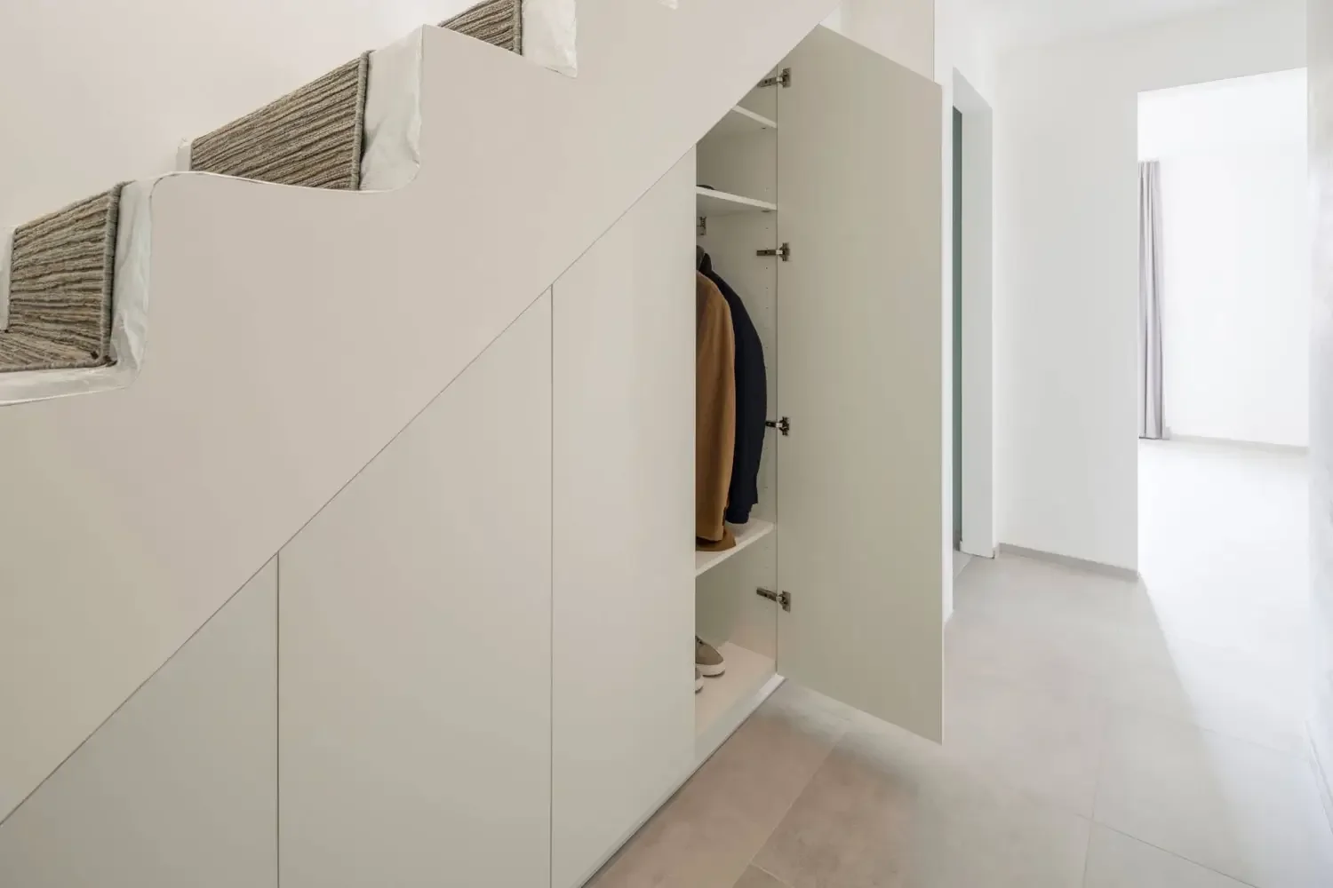 White storage cabinet built under staircase with open door revealing hanging clothes and shoes inside, next to a bright room with white walls and a window with curtains.