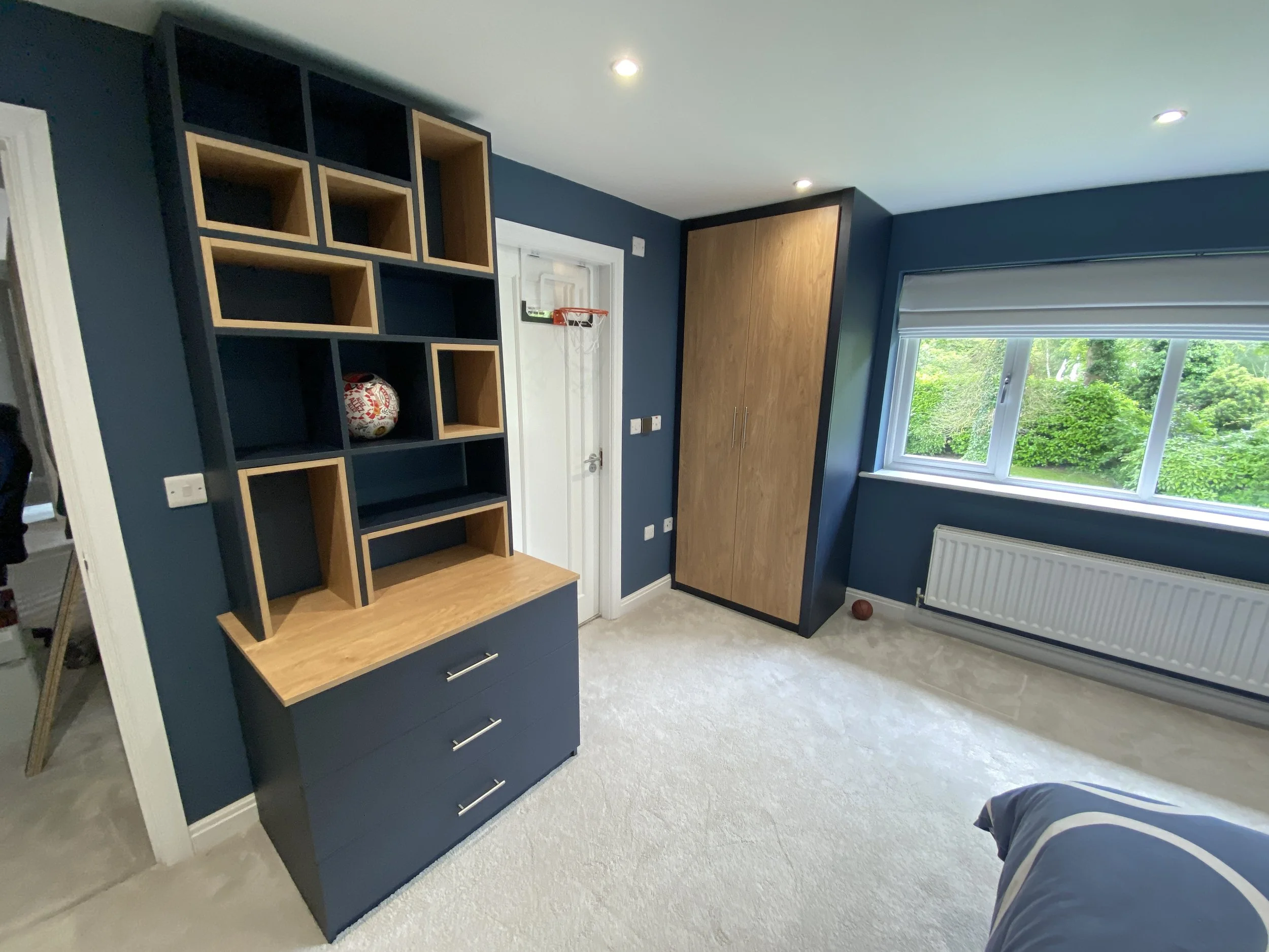 A modern bedroom with a navy blue and wood-tone shelving unit and cabinet, a window with a view of green trees, and beige carpeted flooring.