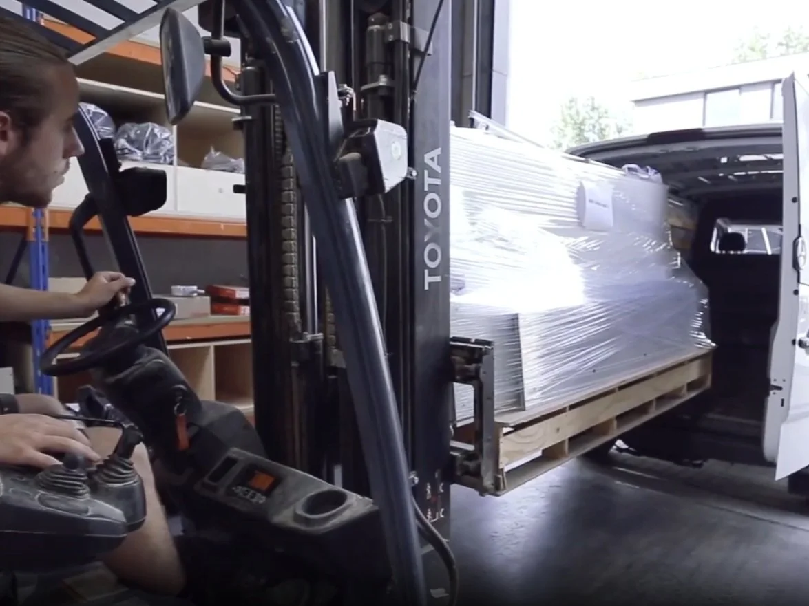 A person operating a forklift truck to unload a large wrapped pallet from the back of a delivery van.