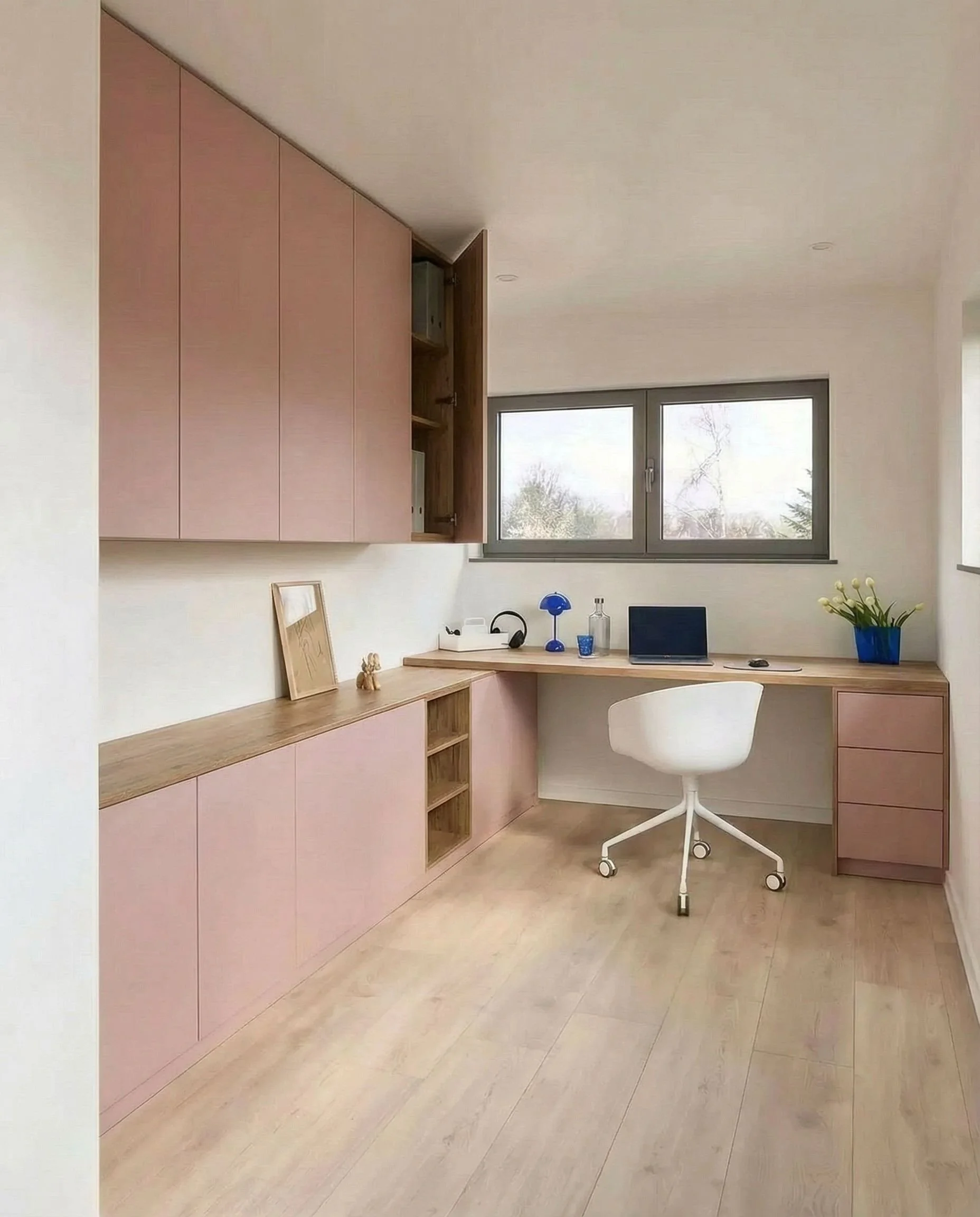 Minimalist home office with light pink and wood cabinetry, a white desk chair, a window, and decorative items including a blue vase with yellow flowers, a blue lamp, and a laptop.