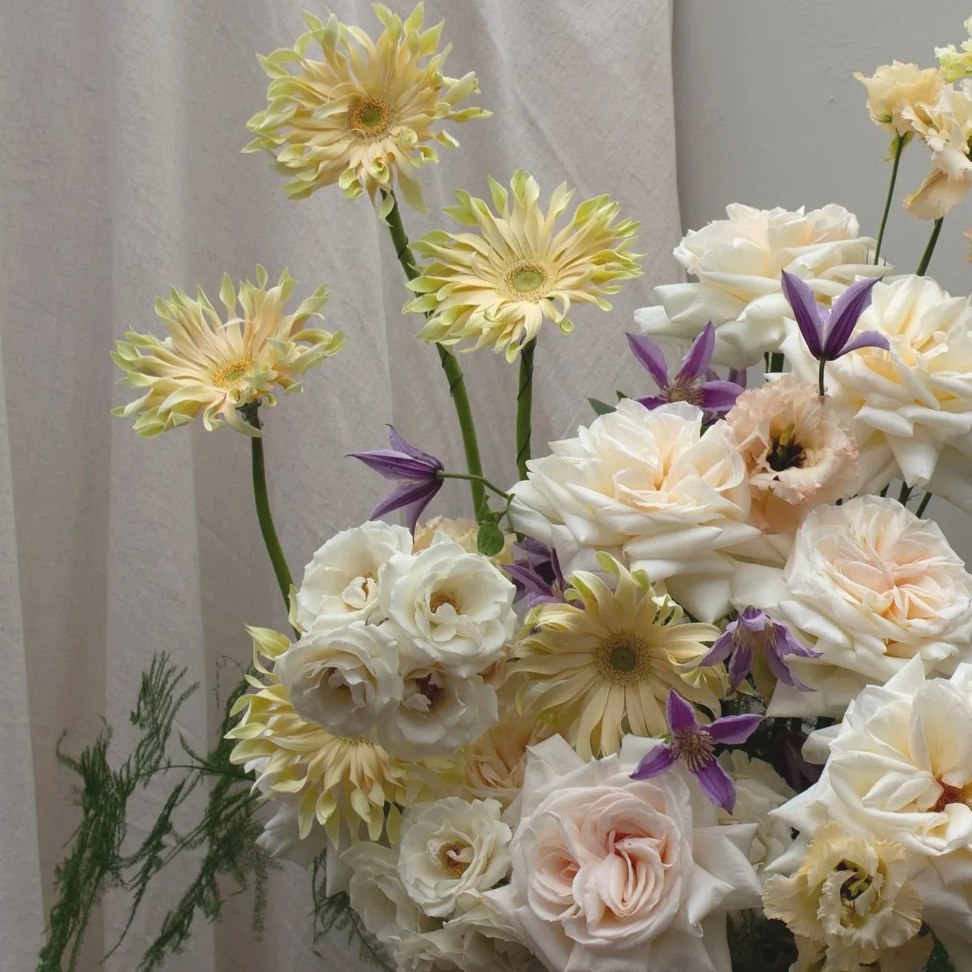 Arrangement of cream, white, and purple flowers, including gerberas, roses, and clematis with green foliage against a neutral background.