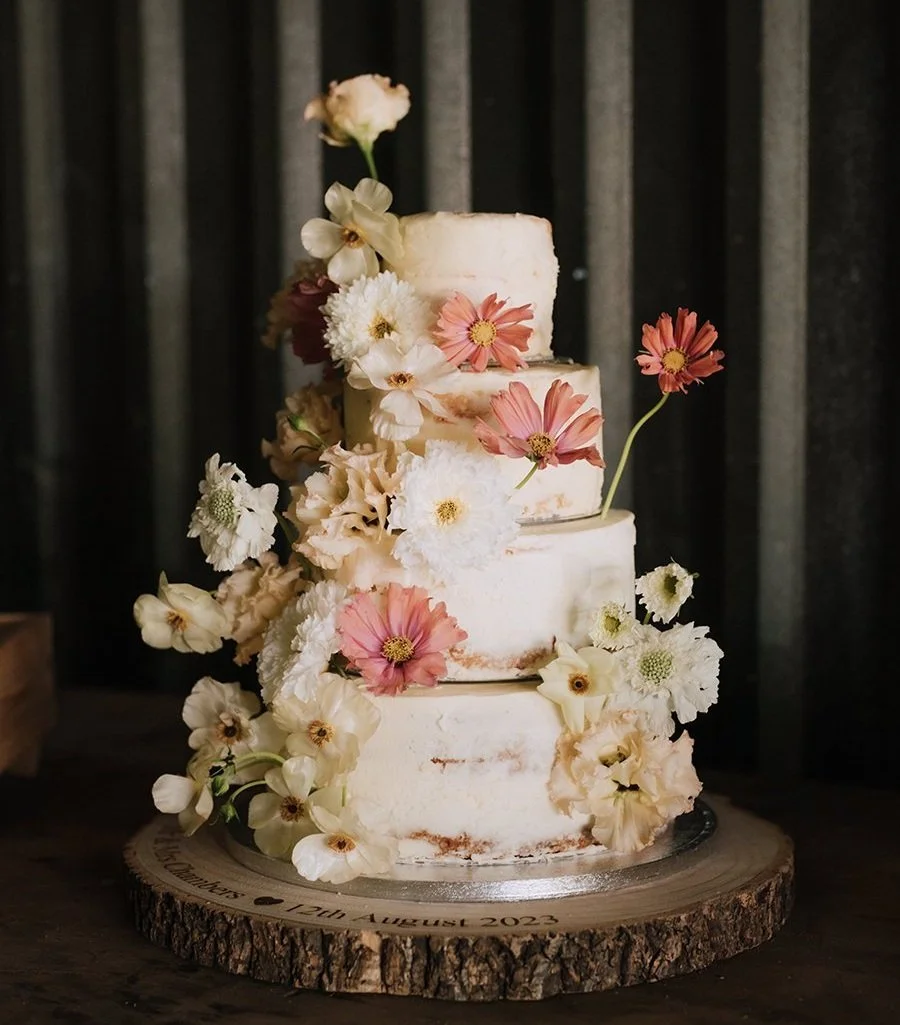 Three-tiered semi-naked wedding cake with pink and white flowers on a wooden base.