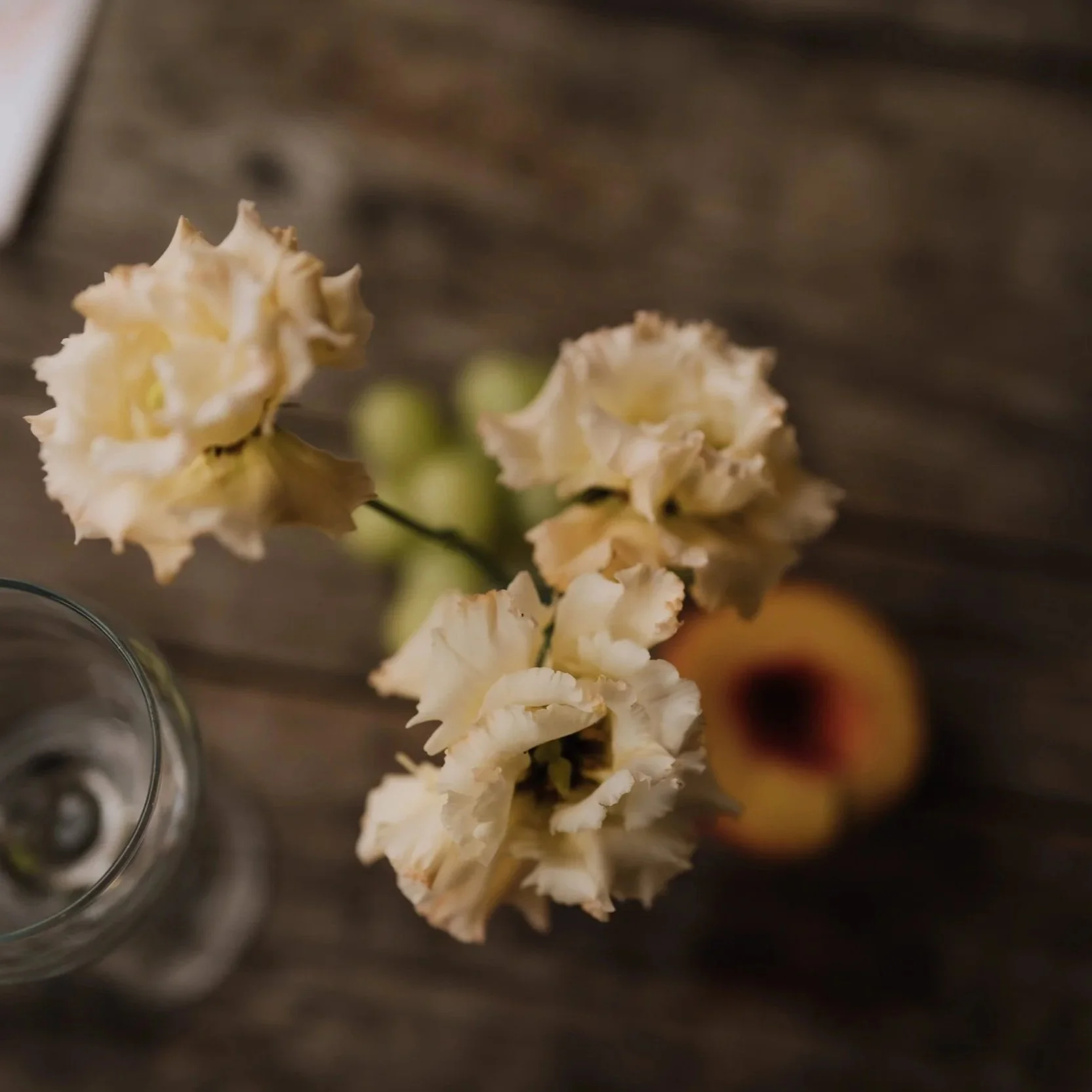 Close-up of a vase with cream-coloured lisianthus, a wine glass, a peach, and some grapes on a rustic wooden table