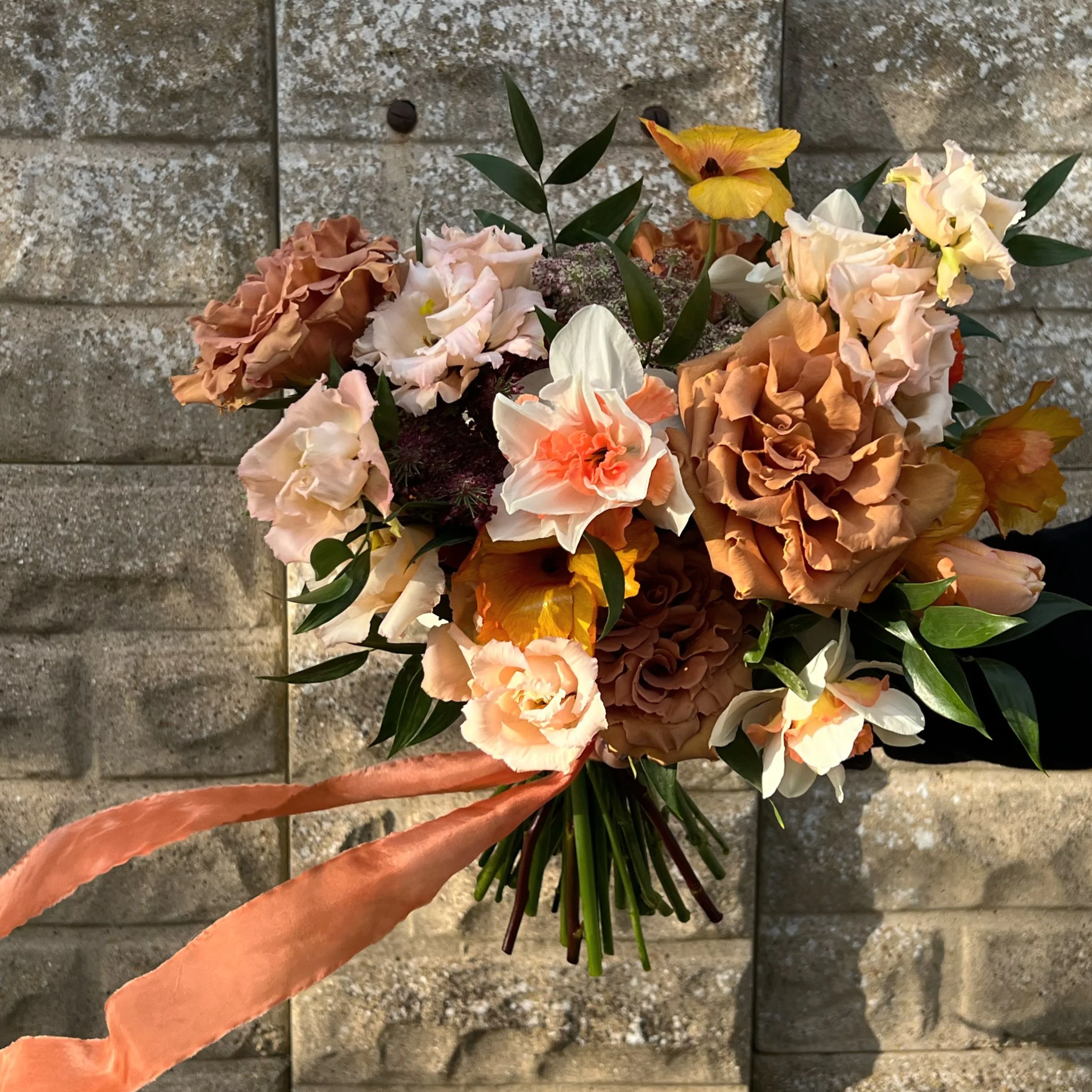 sun kissed spring bridal bouquet, with toffee roses, peach lisianthus, orange butterfly ranunculus and white and orange daffodils, against a stone wall, with a rust silk ribbon