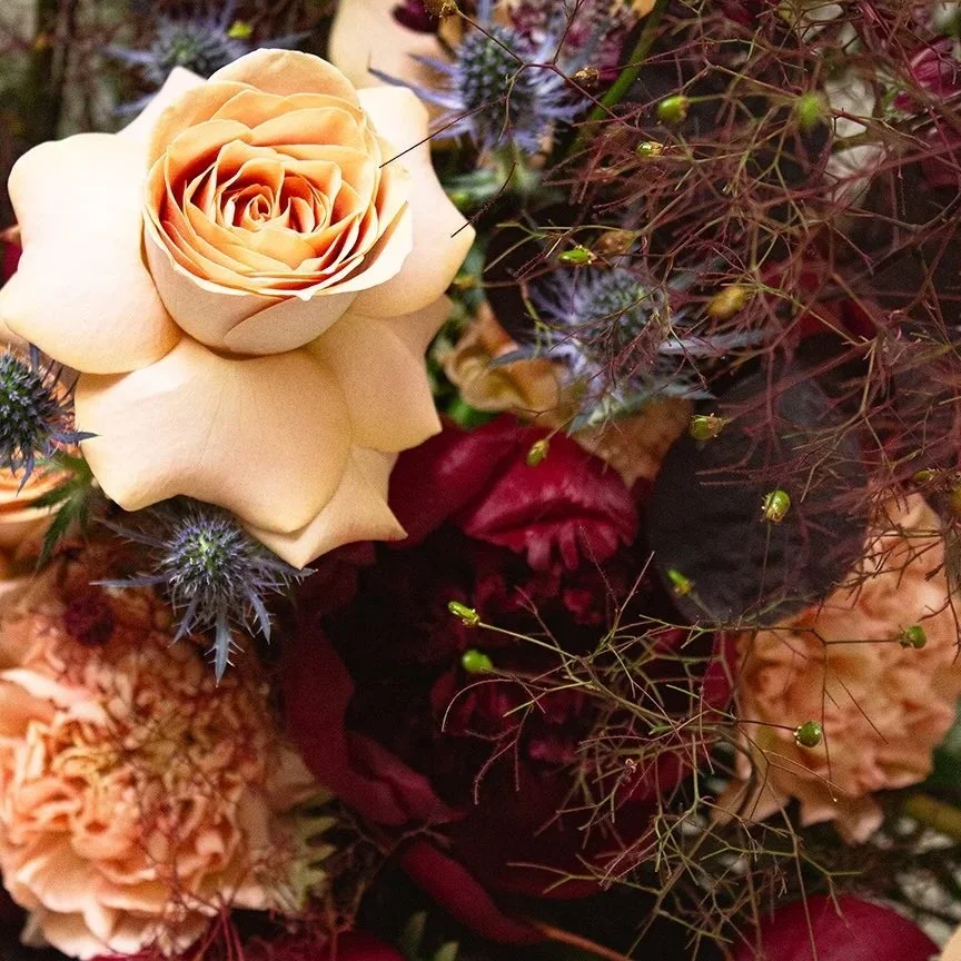 close up of a wedding floral arrangement, featuring roses, carnations, deep red peonies and smokebush