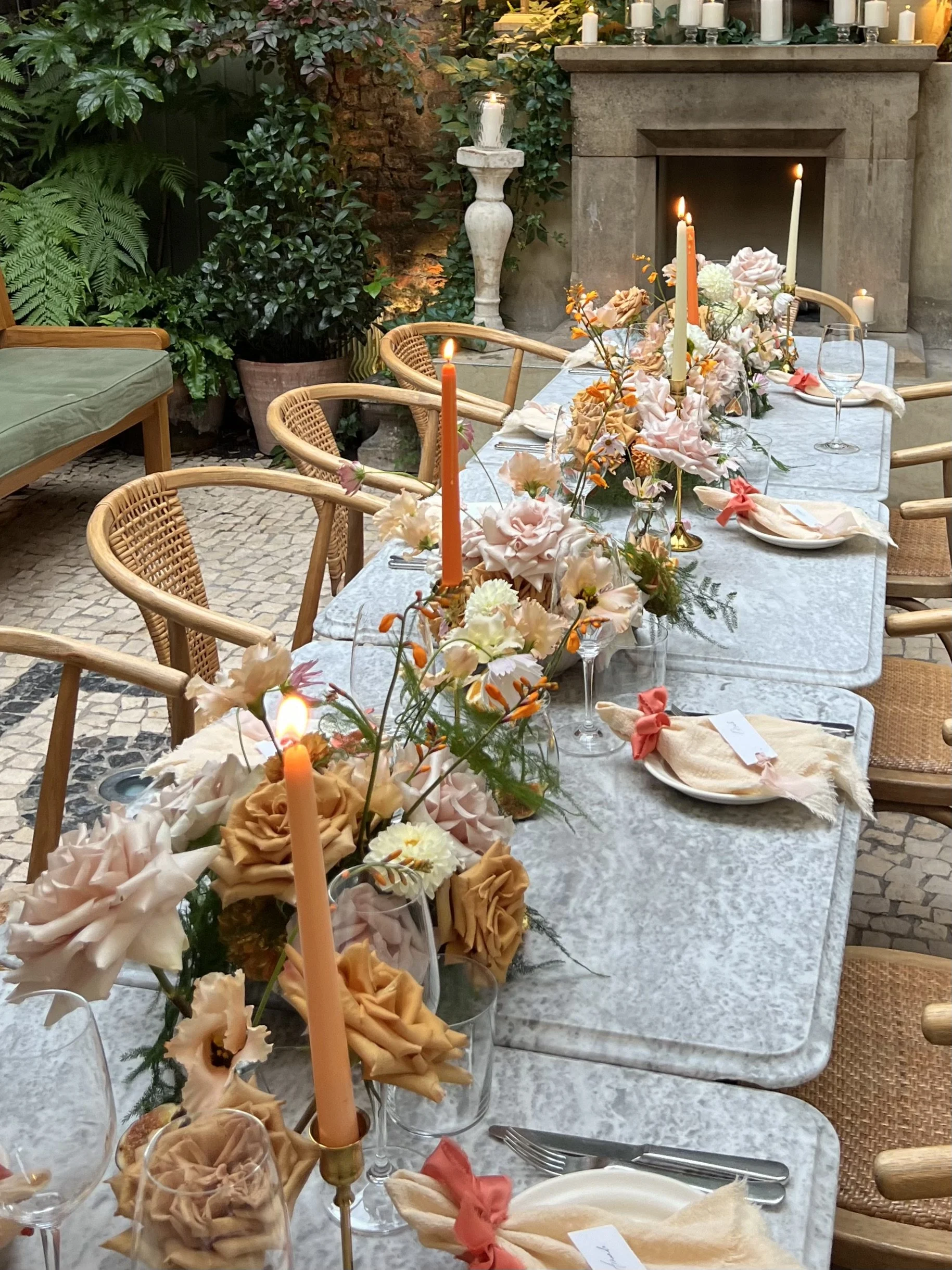 A long dining table decorated with pastel floral arrangements, tall taper candles in gold holders, and place settings with napkins tied with pink ribbons. The table is set in an outdoor garden area with greenery and stone walls.