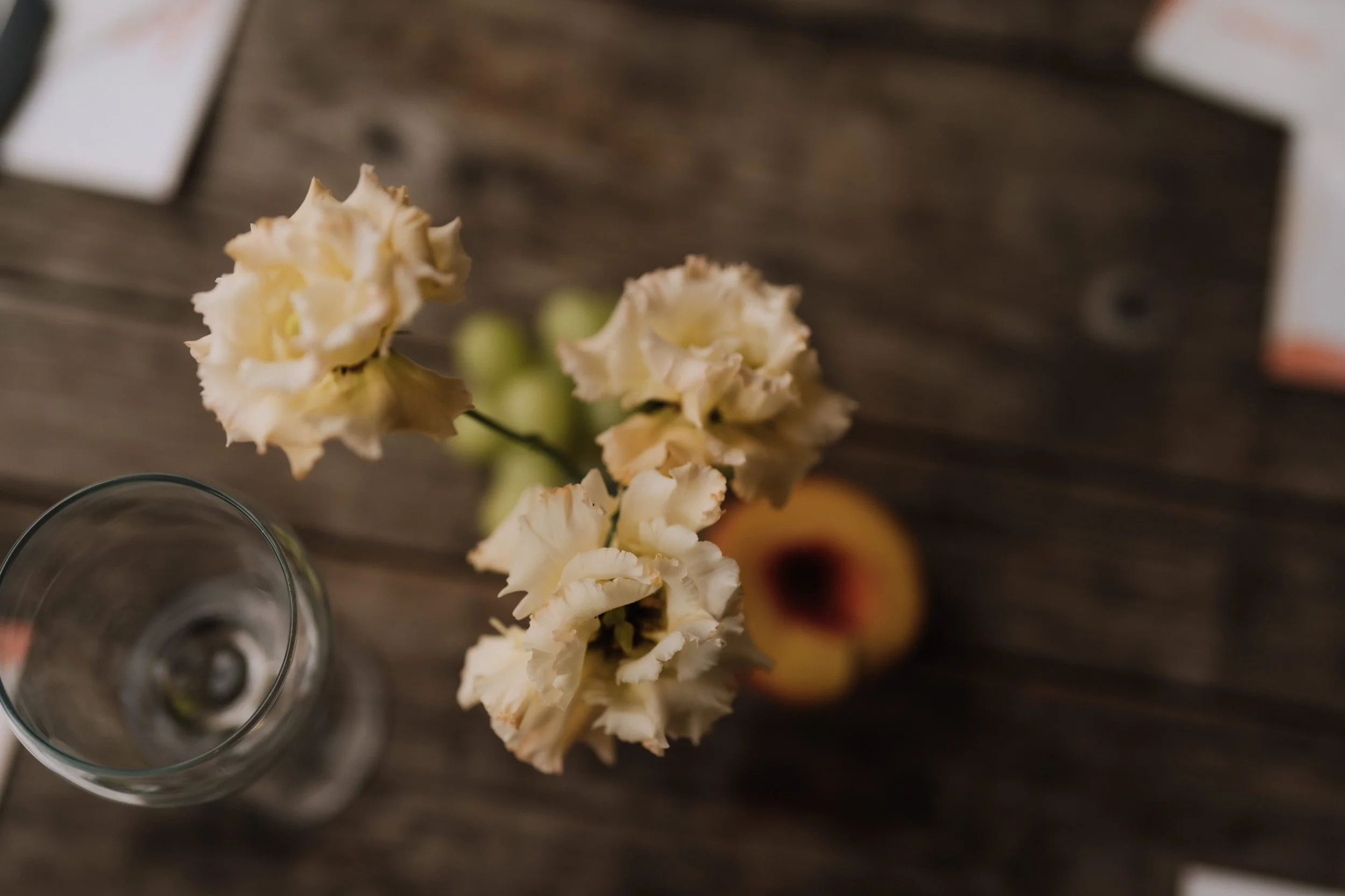 Close-up of a vase with cream-coloured lisianthus, a wine glass, a peach, and some grapes on a rustic wooden table
