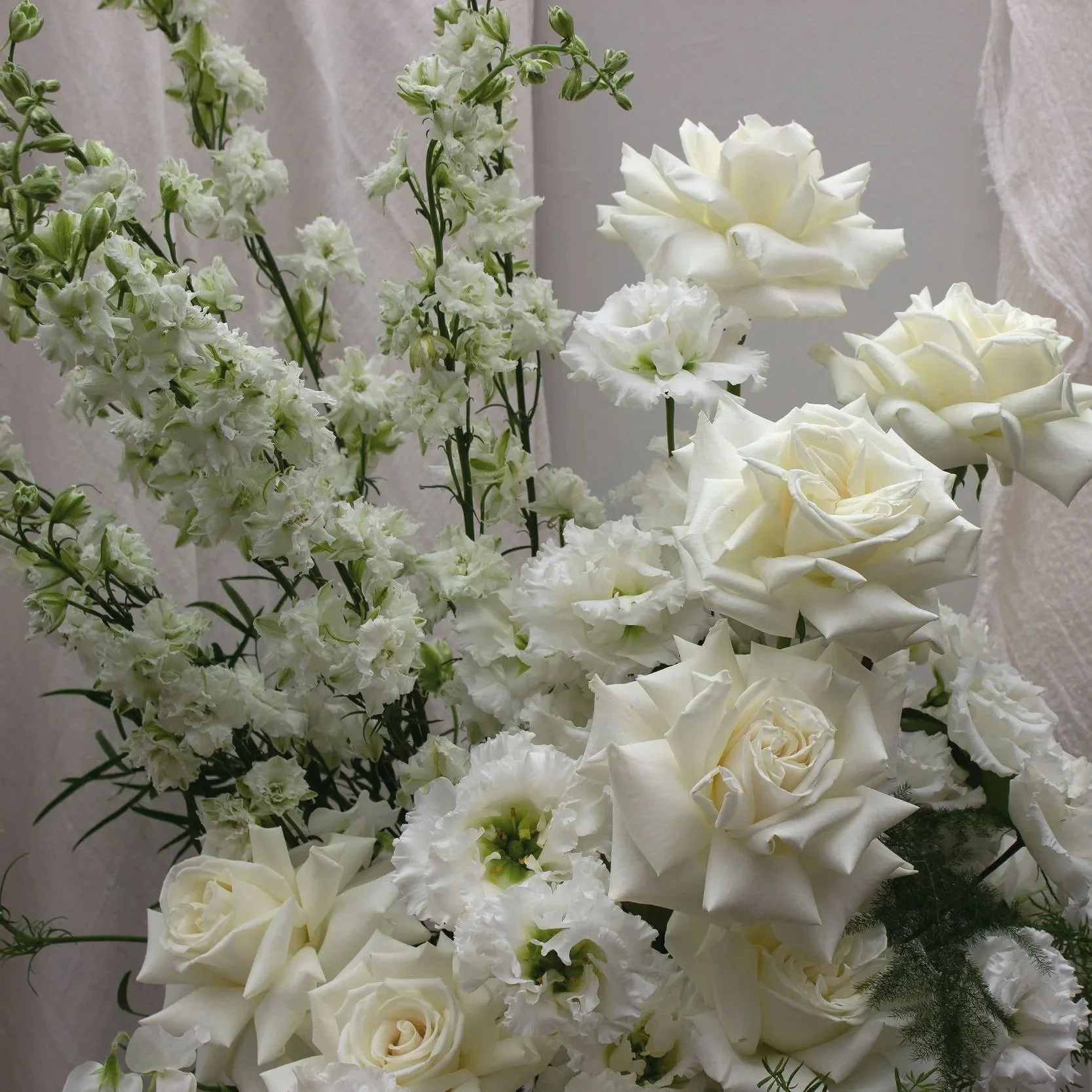A close-up of a floral arrangement featuring white roses and white delphiniums, with a soft fabric background.