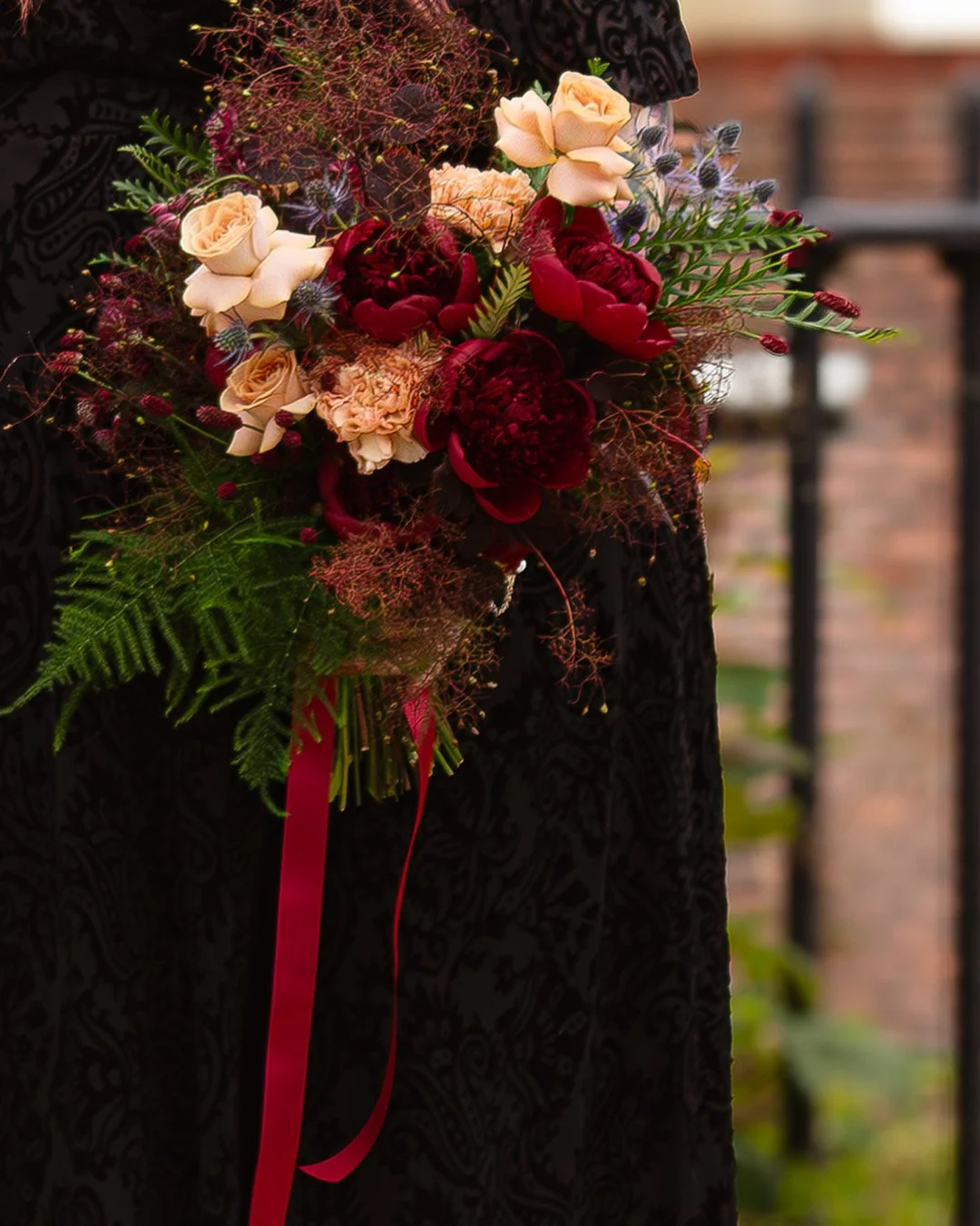 Close-up of a person holding a bouquet of flowers featuring cream roses, dark red peonies, and various greenery, attached with a red ribbon. The person is wearing a black embossed dress with an elaborate pattern.